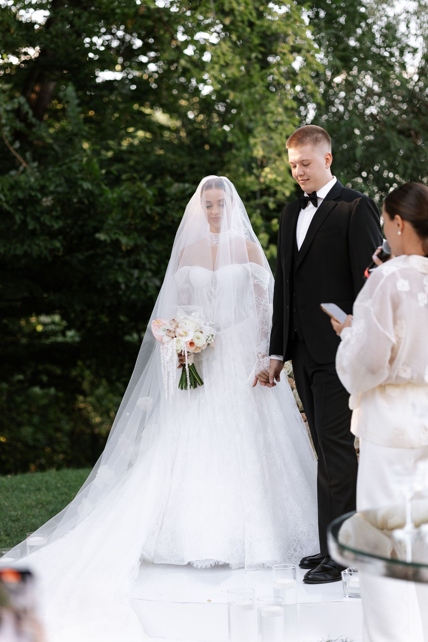 A bride in a flowing white gown and veil walks with her wedding party outdoors.