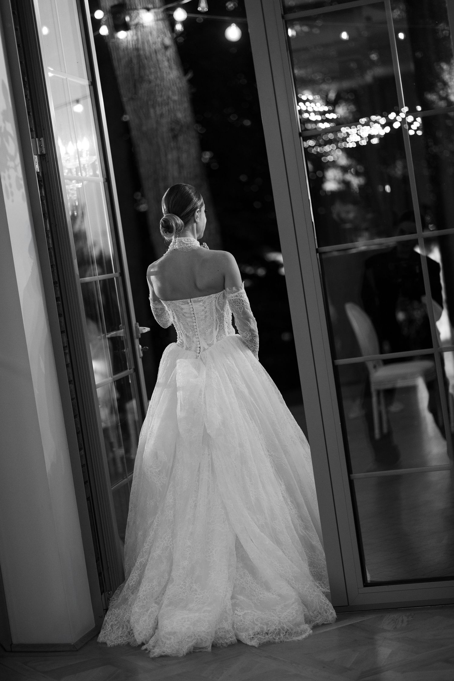 A bride in a flowing ballgown walks through a dimly lit venue hallway in black and white photography.