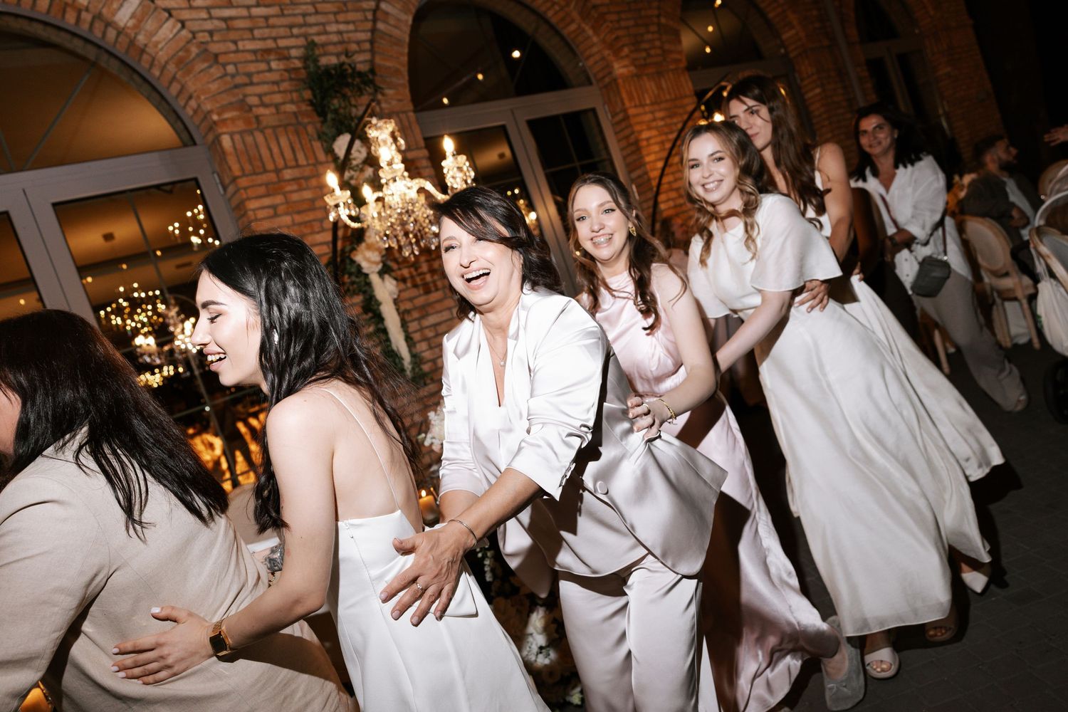 Wedding guests dance and celebrate on a dance floor under brick arches and chandeliers.