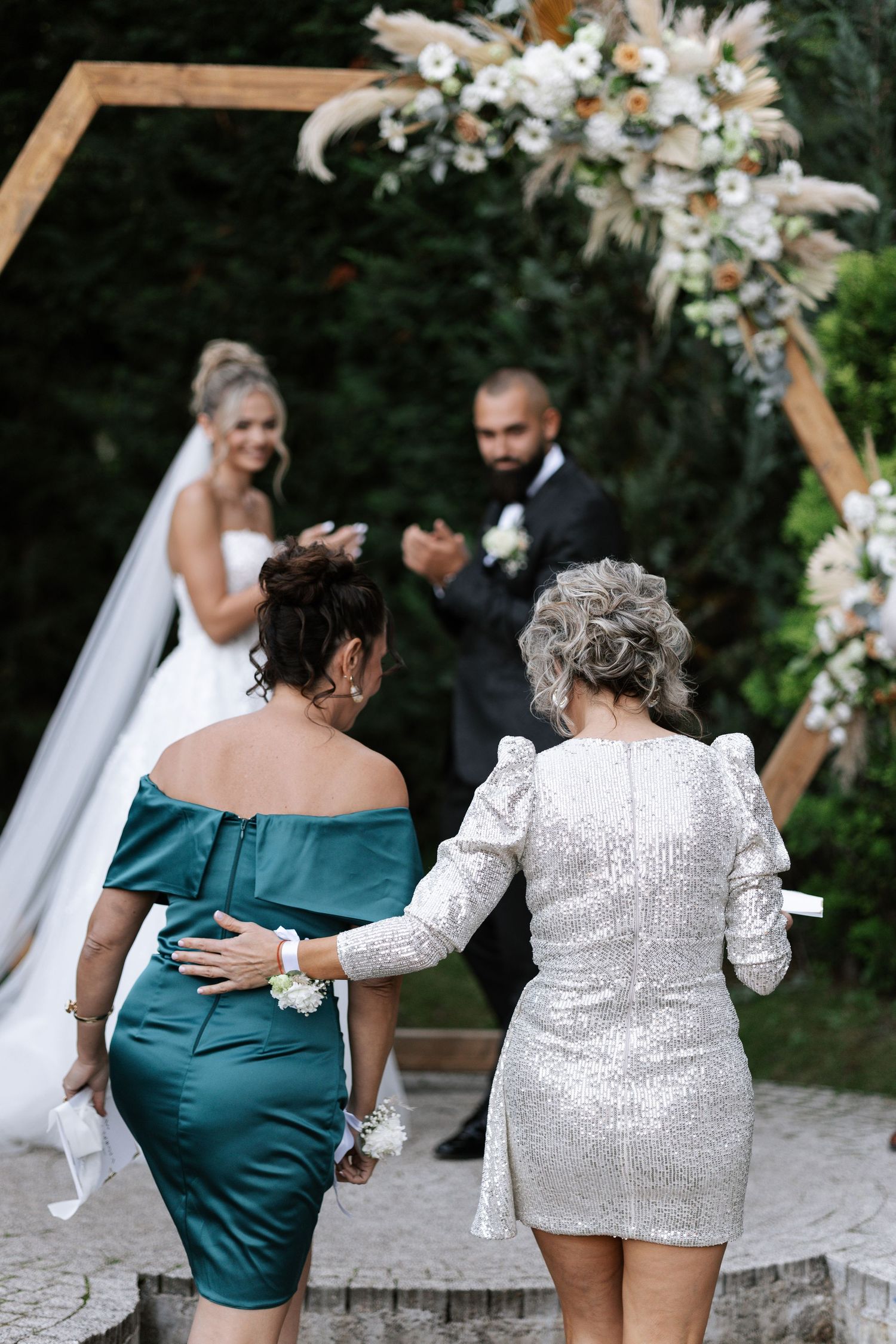 Two wedding guests in elegant dresses walk towards a floral arch ceremony setup outdoors.