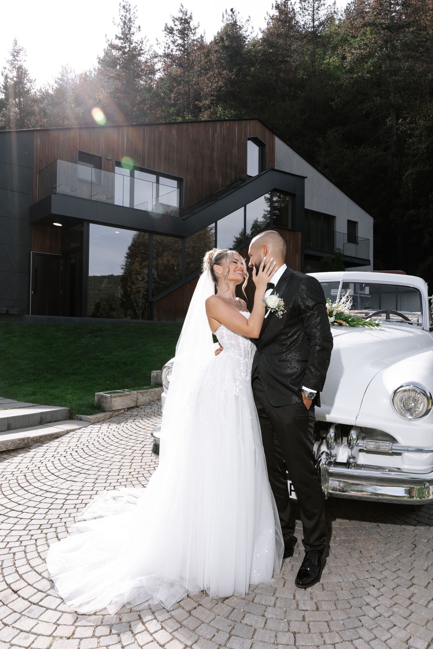Wedding couple sharing romantic kisses in front of vintage car and modern building exterior.