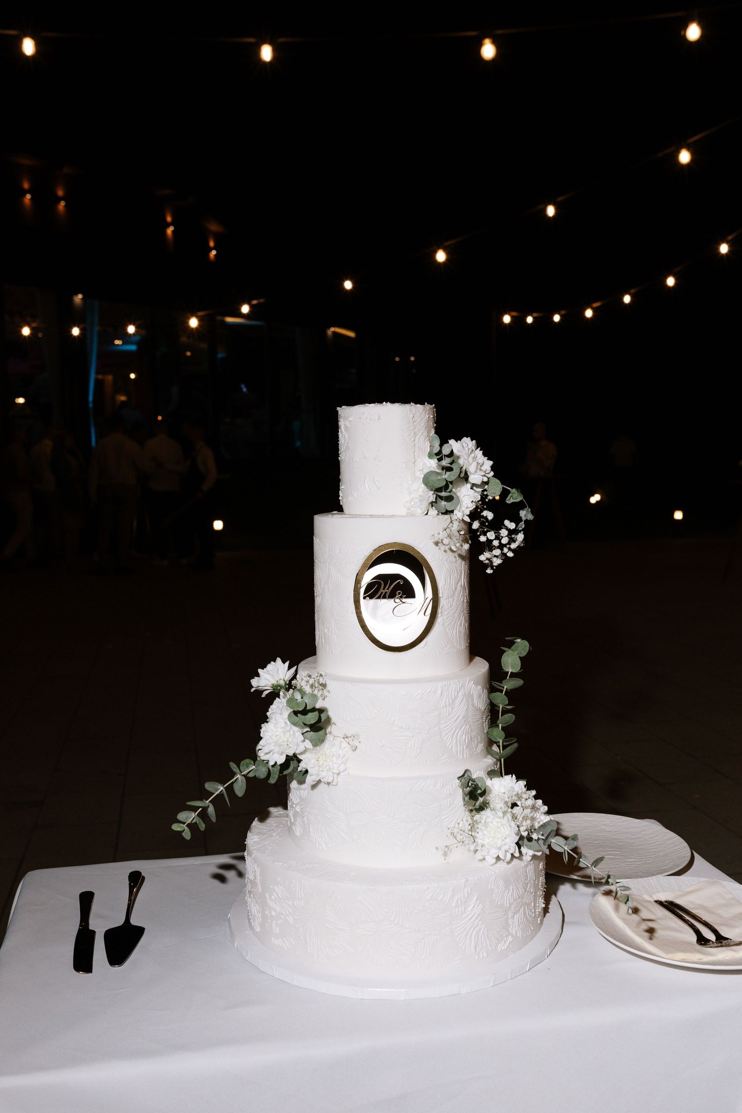 A white tiered wedding cake with floral decorations displayed under twinkling lights at a reception.