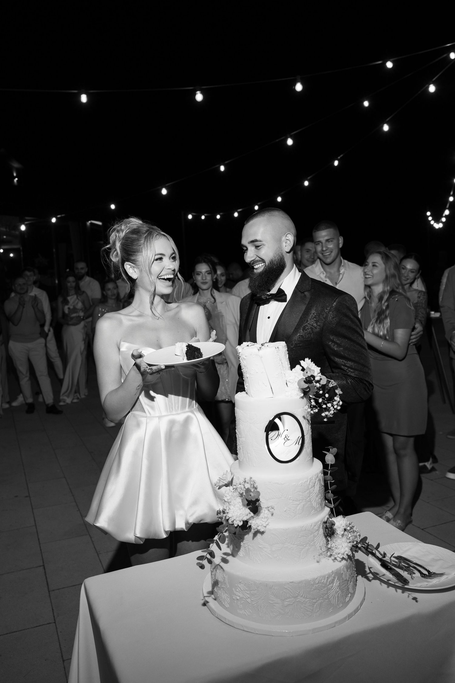 Black and white photo of a wedding couple sharing a joyful moment while cutting their elegant tiered cake under string lights.