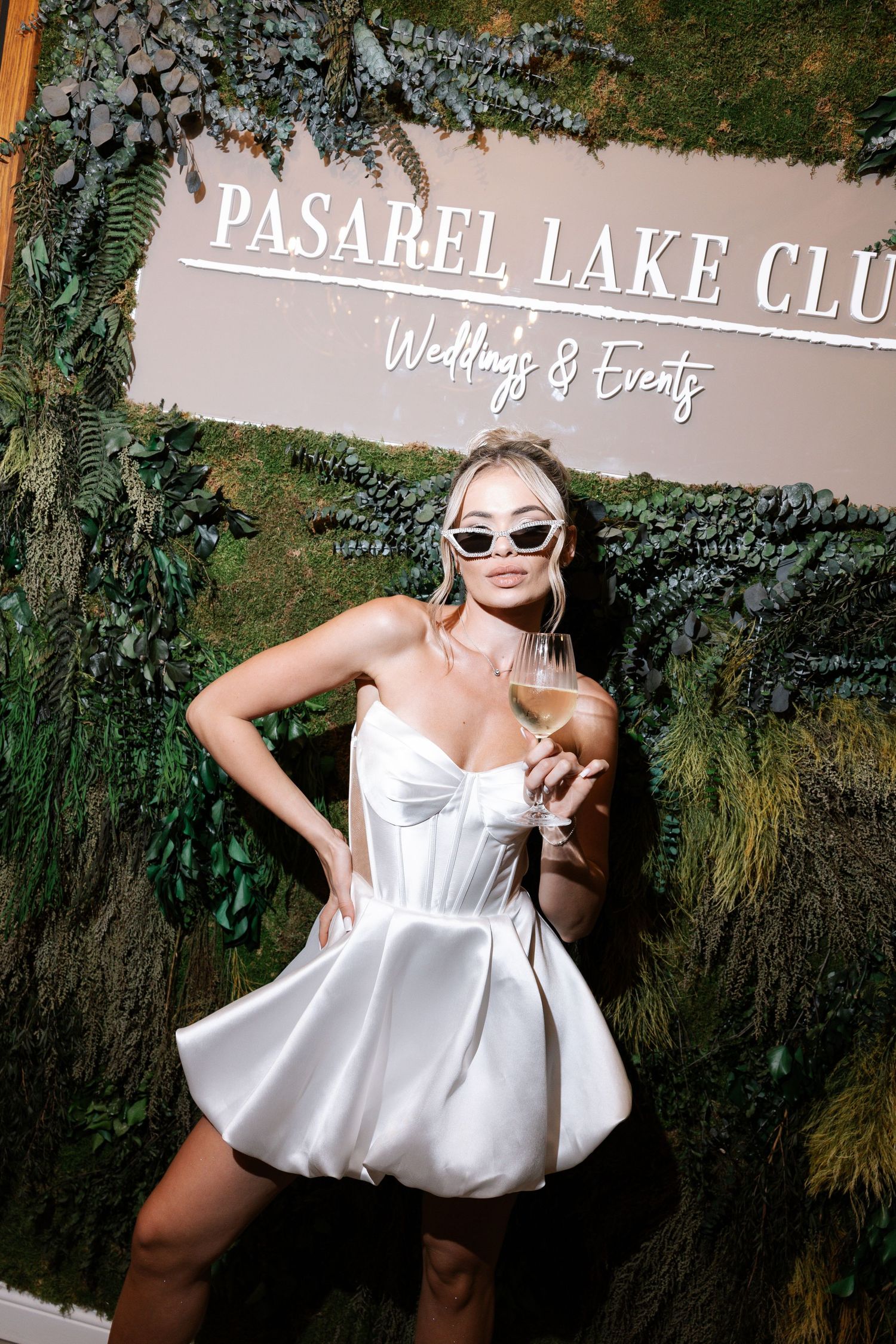 A person in a white dress poses playfully in front of a hedge wall with event venue signage.