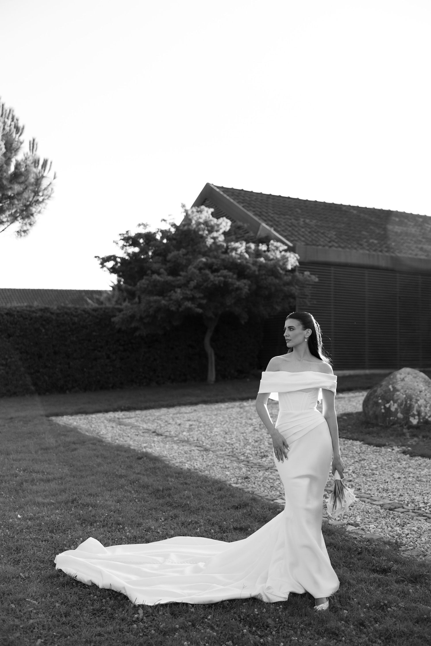 A bride in an elegant white gown with a long flowing train poses gracefully on a grassy lawn in black and white.