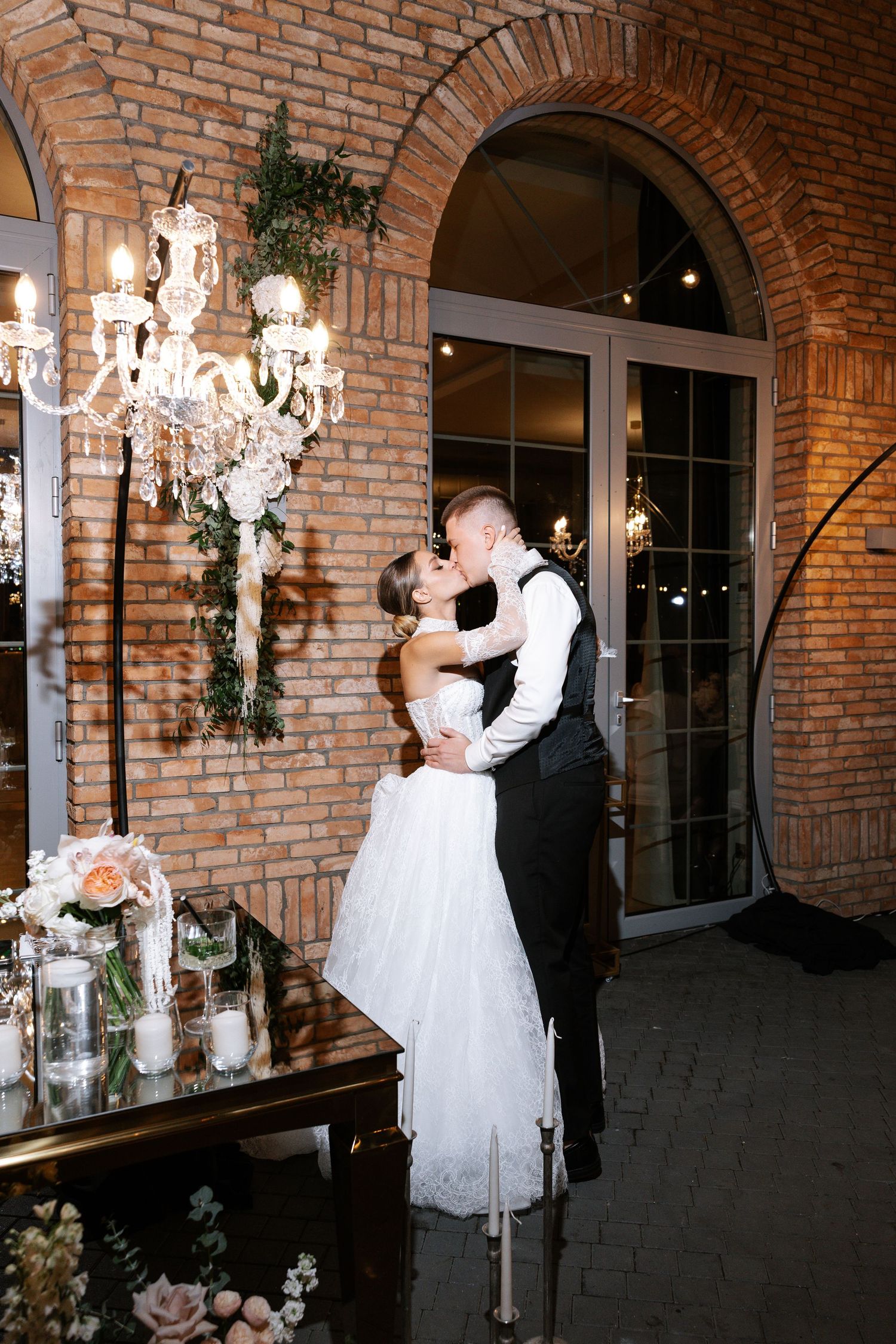 Romantic wedding couple embraces under crystal chandelier and exposed brick archway at elegant indoor venue.