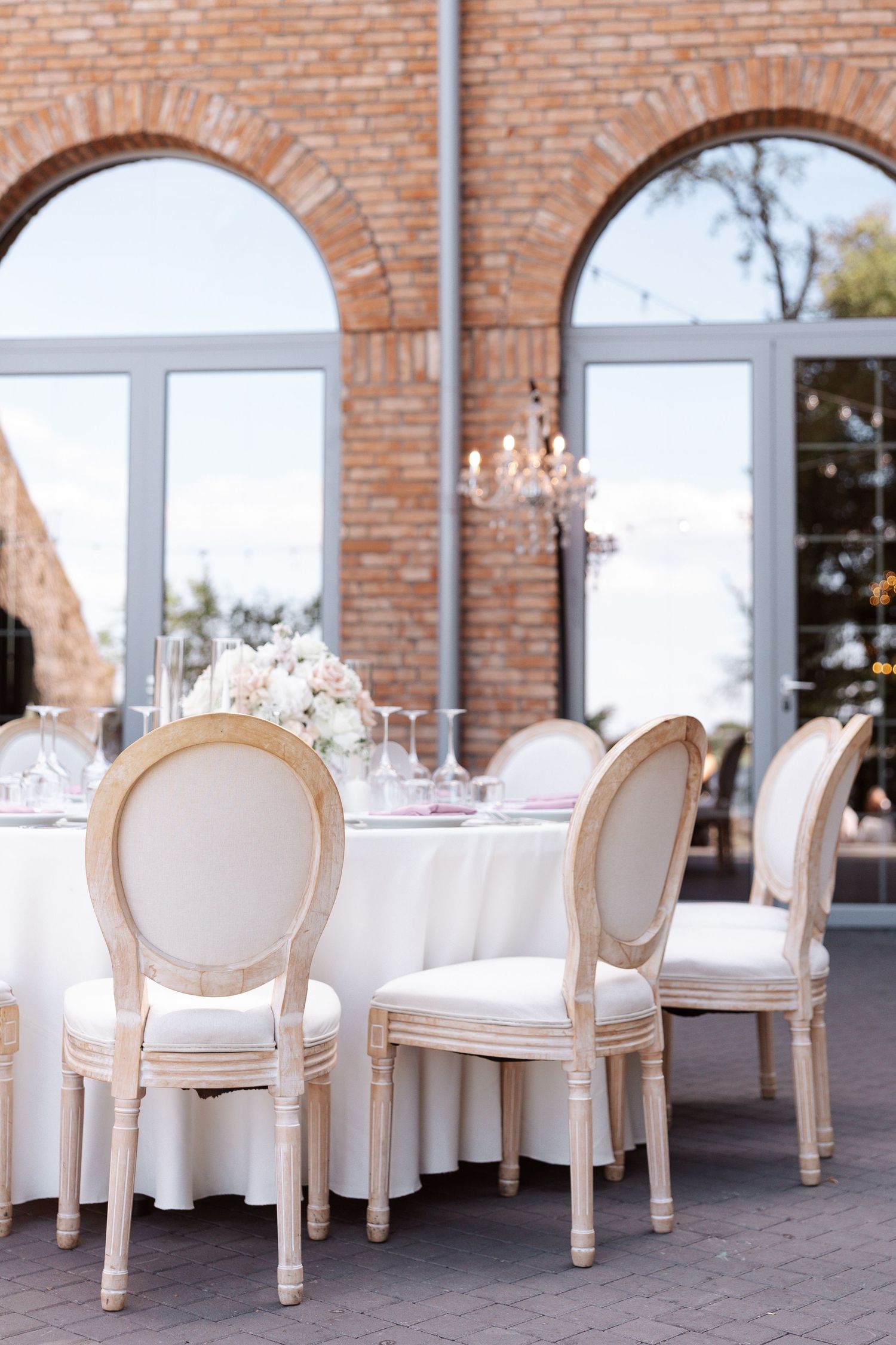 Elegant wedding reception table setting with white chairs and crystal chandelier in a brick-walled venue.