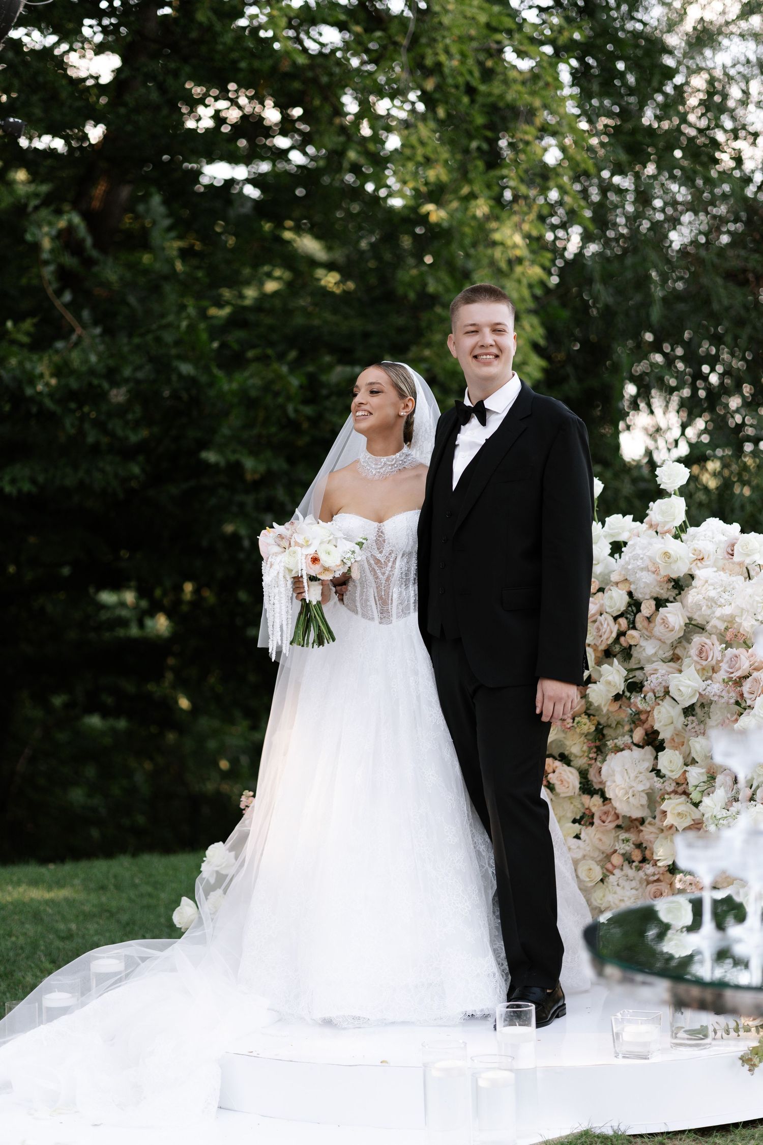 A wedding couple stands at their outdoor ceremony altar surrounded by elegant white floral arrangements.