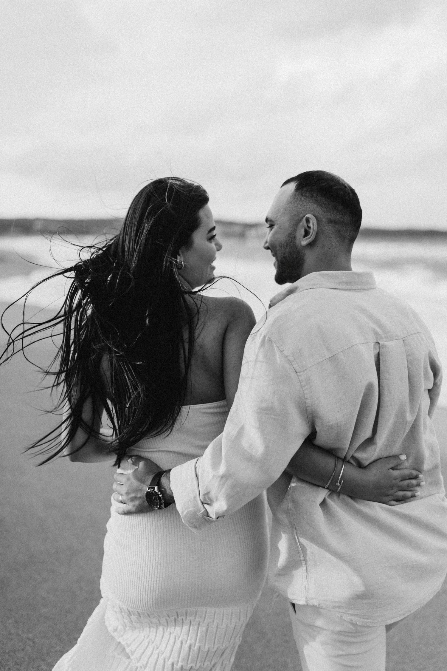 A couple in white outfits embracing on a windy beach with their hair flowing in the breeze.