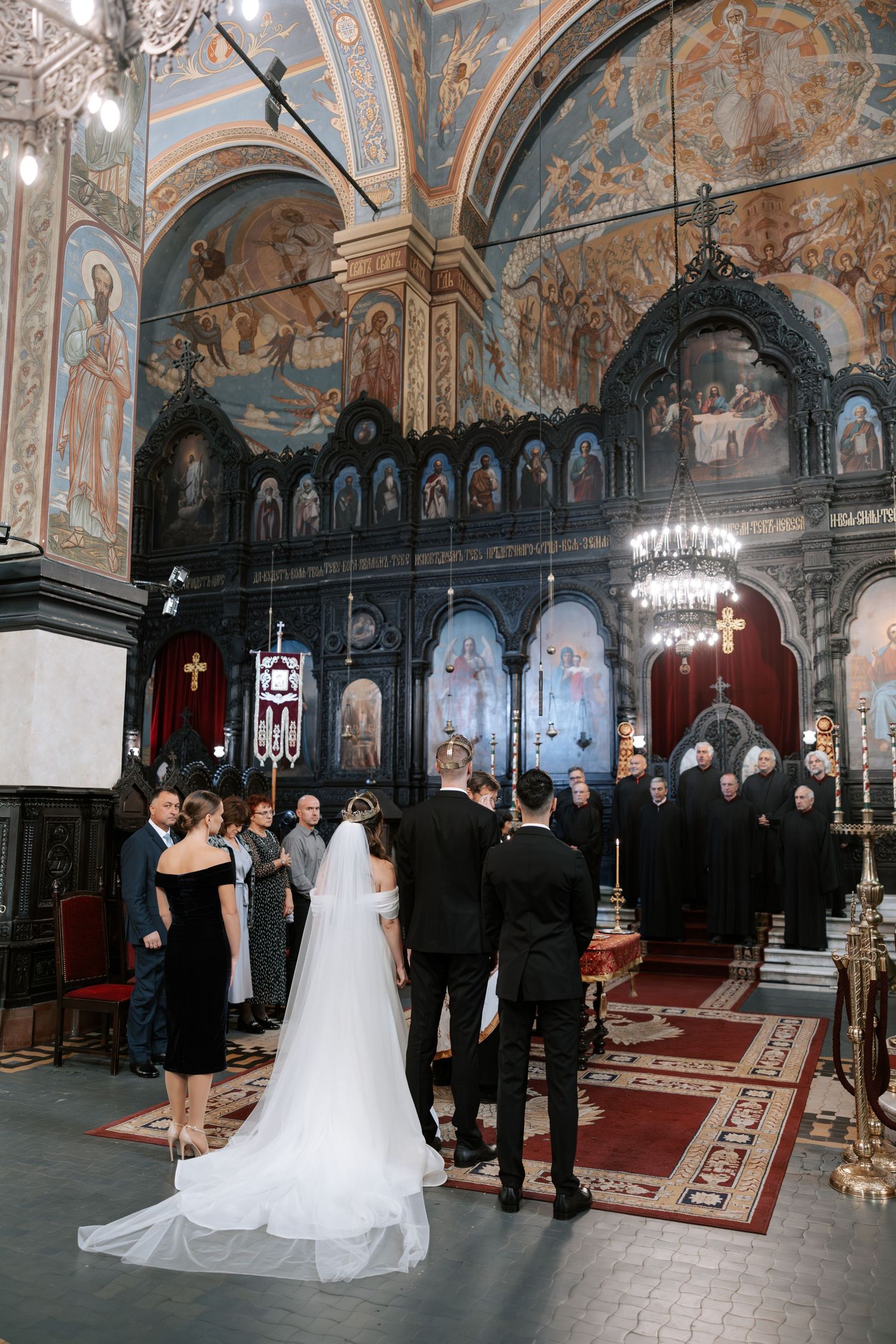 A wedding ceremony takes place inside an ornate Orthodox church with beautiful frescoed walls and crystal chandeliers.