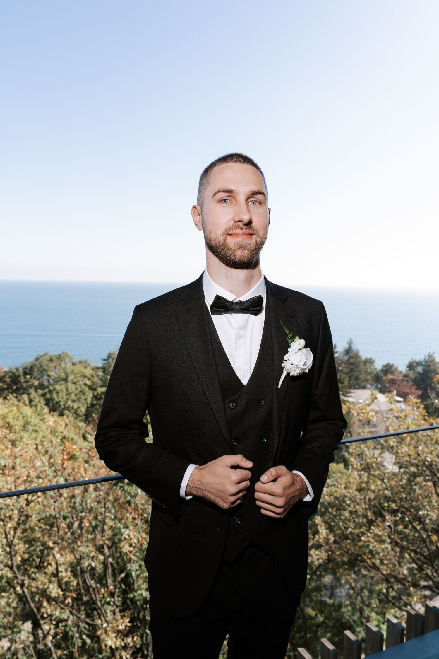 Groom in classic black tuxedo poses against ocean view backdrop.