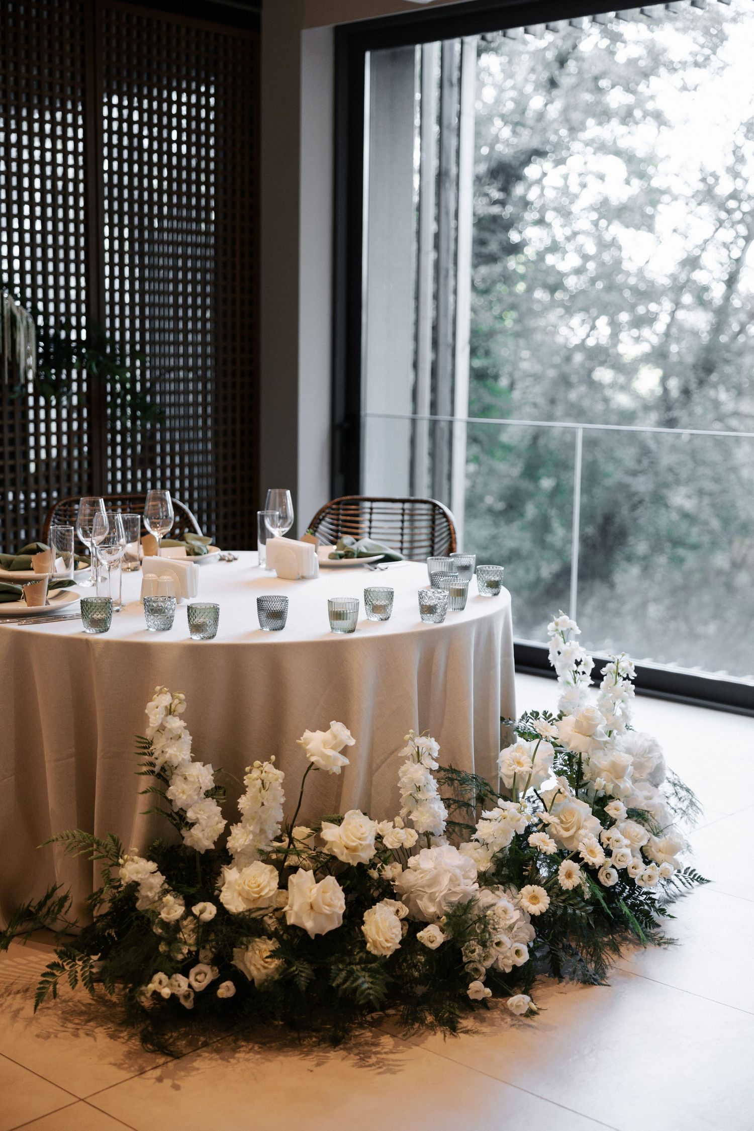 Round table with white tablecloth adorned with lush white floral arrangements near floor-to-ceiling windows.