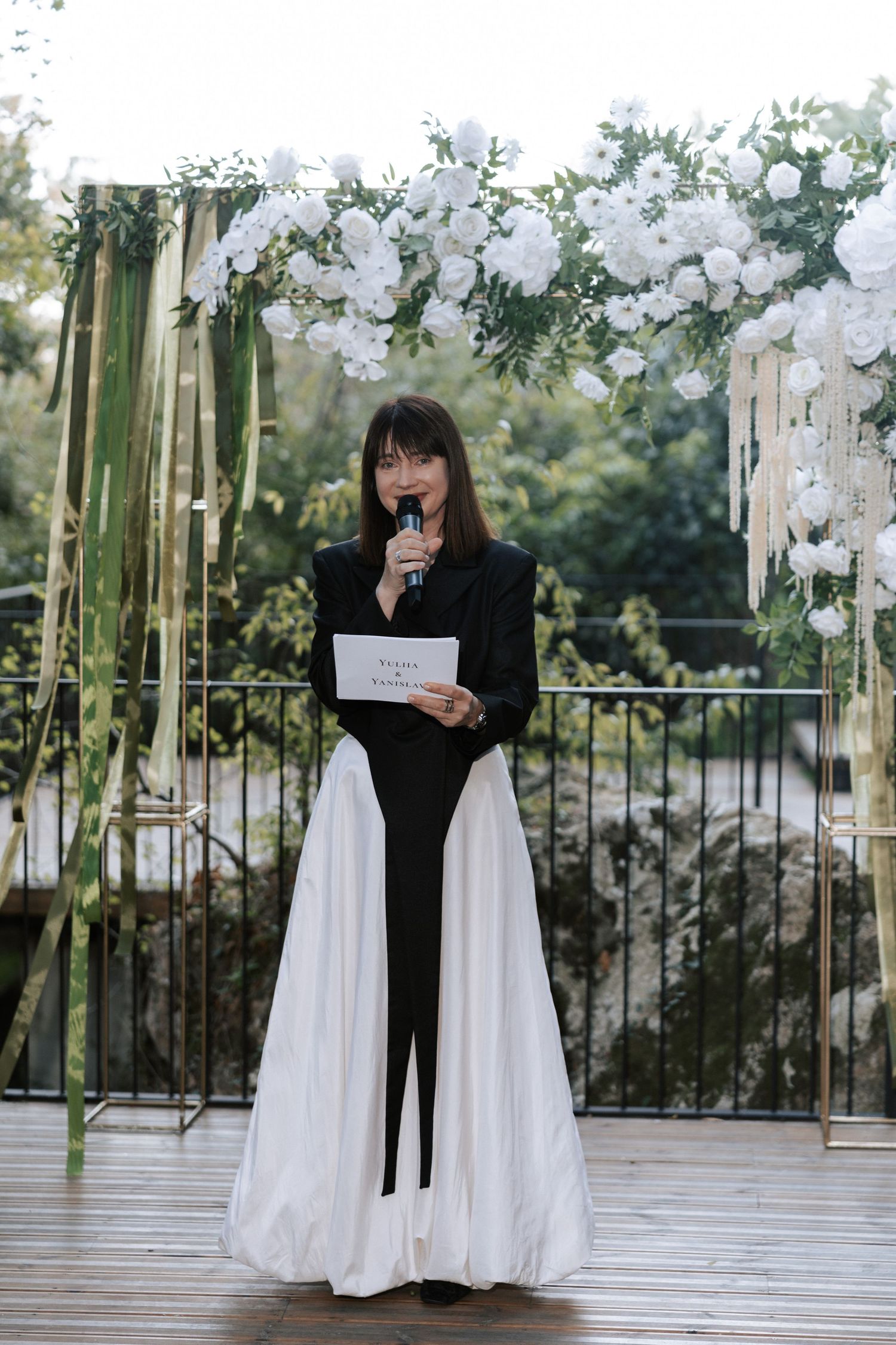 An officiant in black and white stands beneath a floral arch during an outdoor ceremony.
