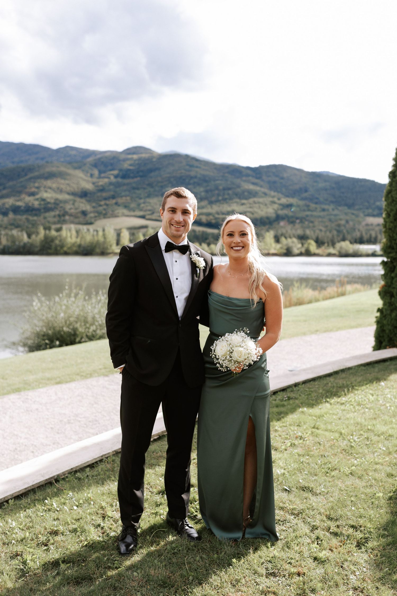 Couple in formal attire pose on lawn with mountain lake backdrop.Wedding photographer Bulgaria