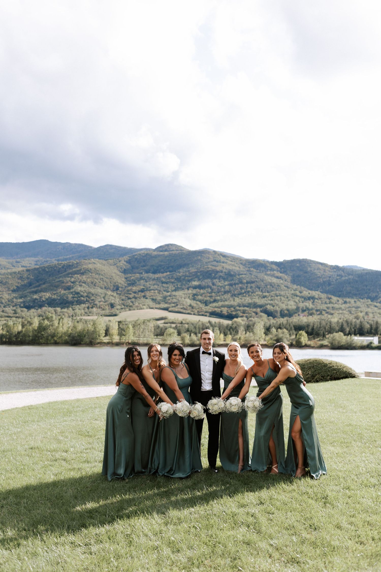 Bridal party in sage green dresses poses on lawn with mountain and lake backdrop.