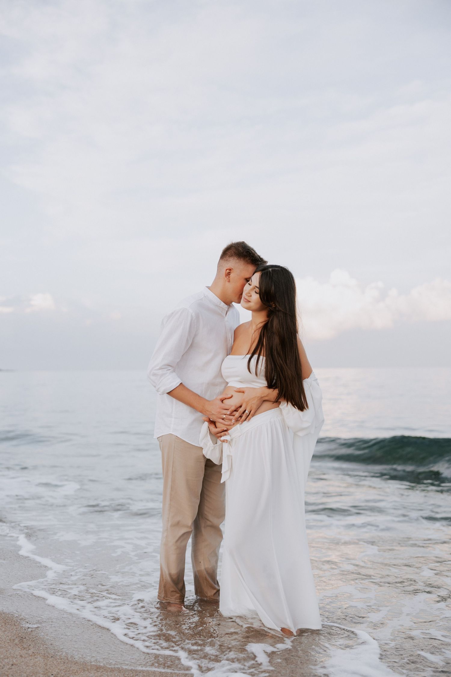 A couple stands in gentle waves during their beach maternity photo session. Family photographer Burgas Bulgaria