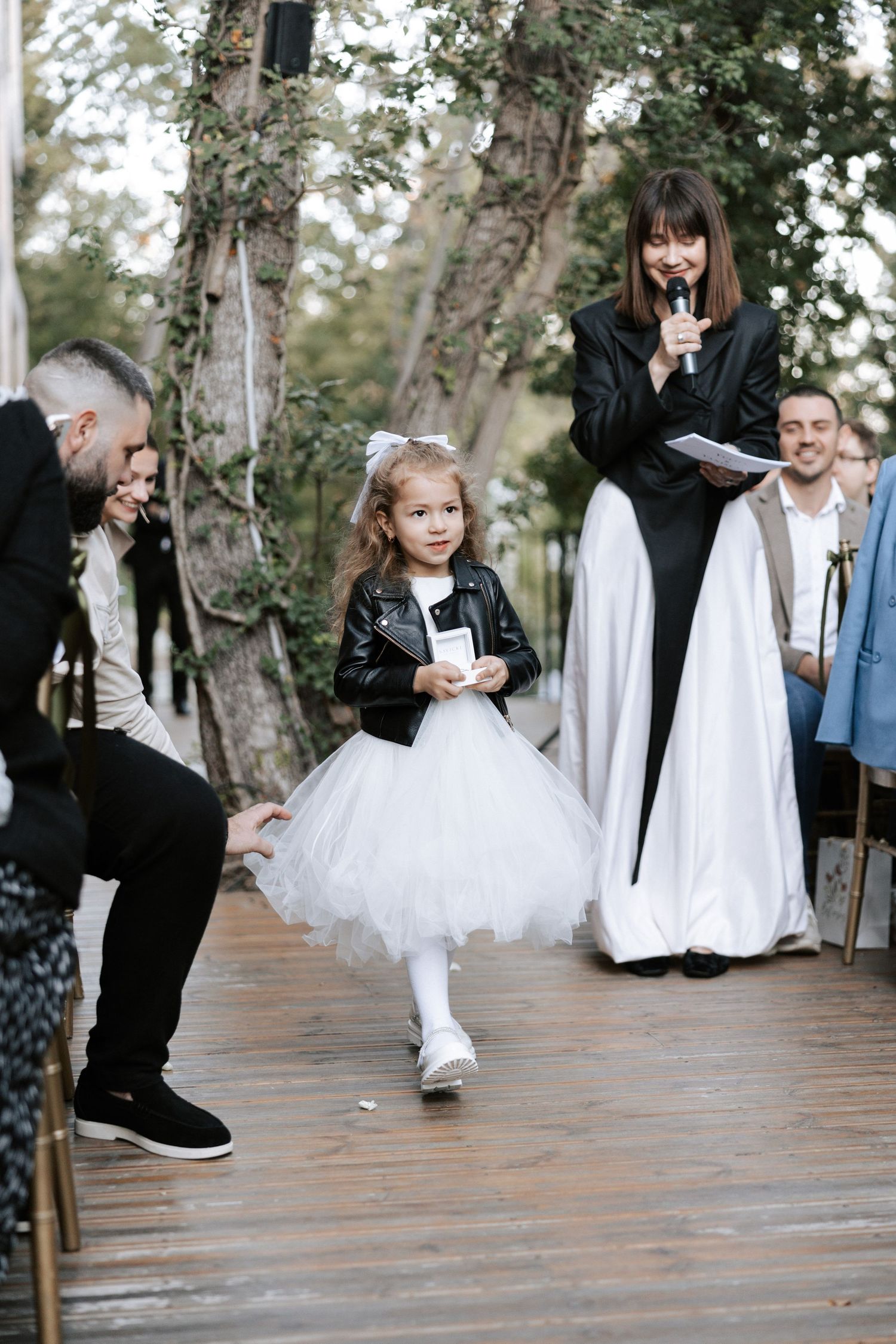 A flower girl in white tulle dress and black leather jacket walks during wedding ceremony.