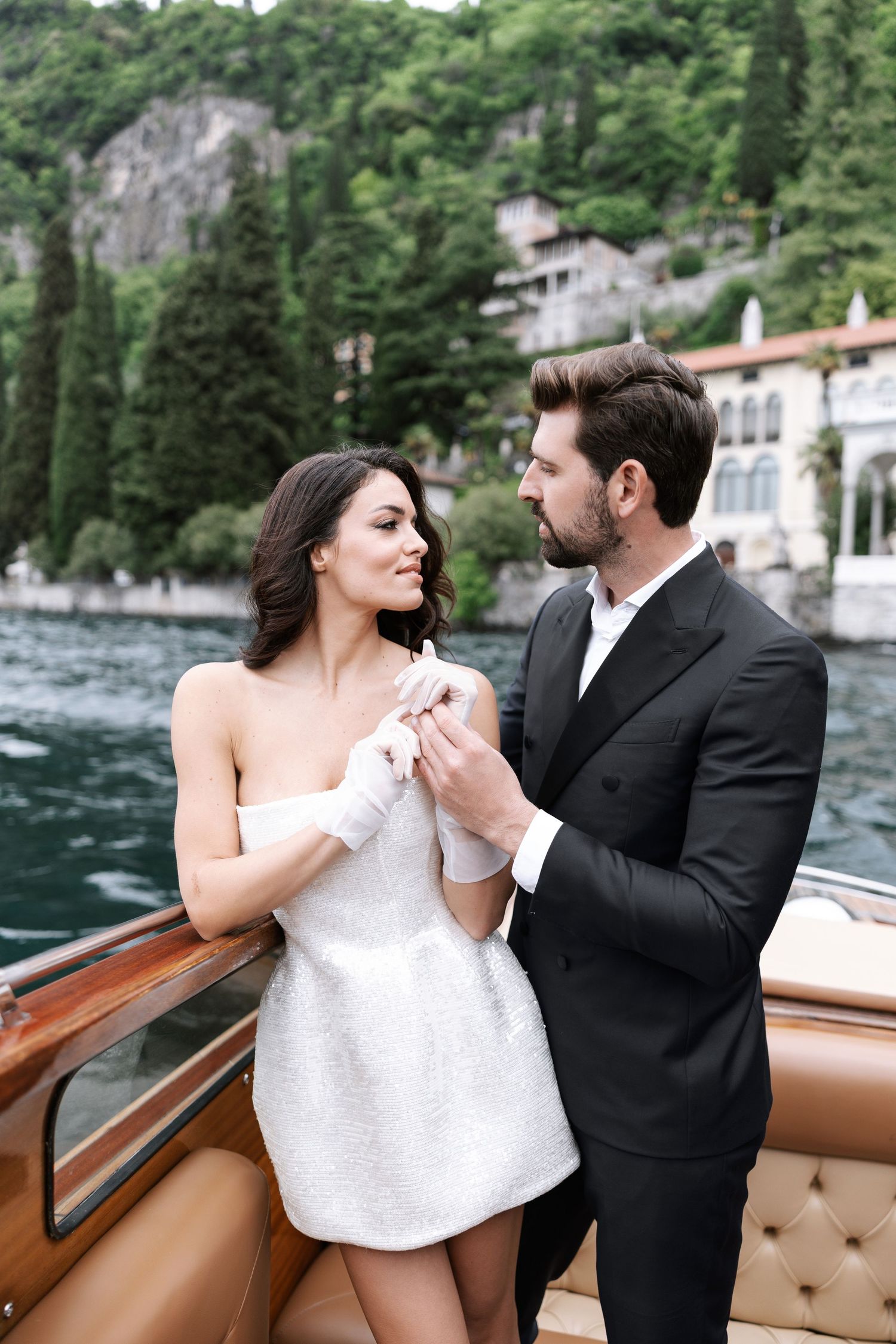 A romantic couple in formal attire share an intimate moment on a luxury boat with Lake Como's scenic shoreline in the background.