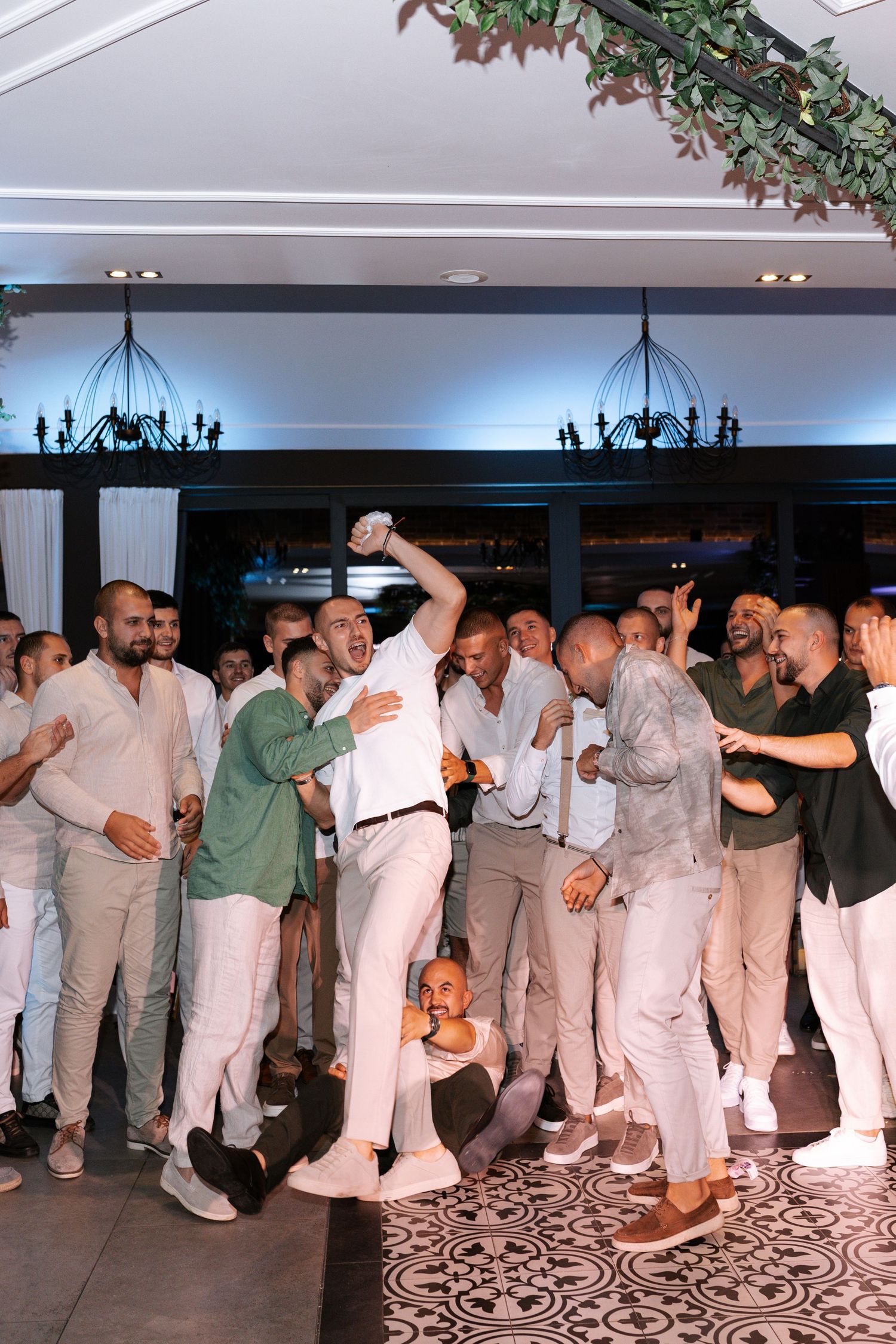 Wedding guests in white and beige attire dance together in a line formation at an indoor reception venue.