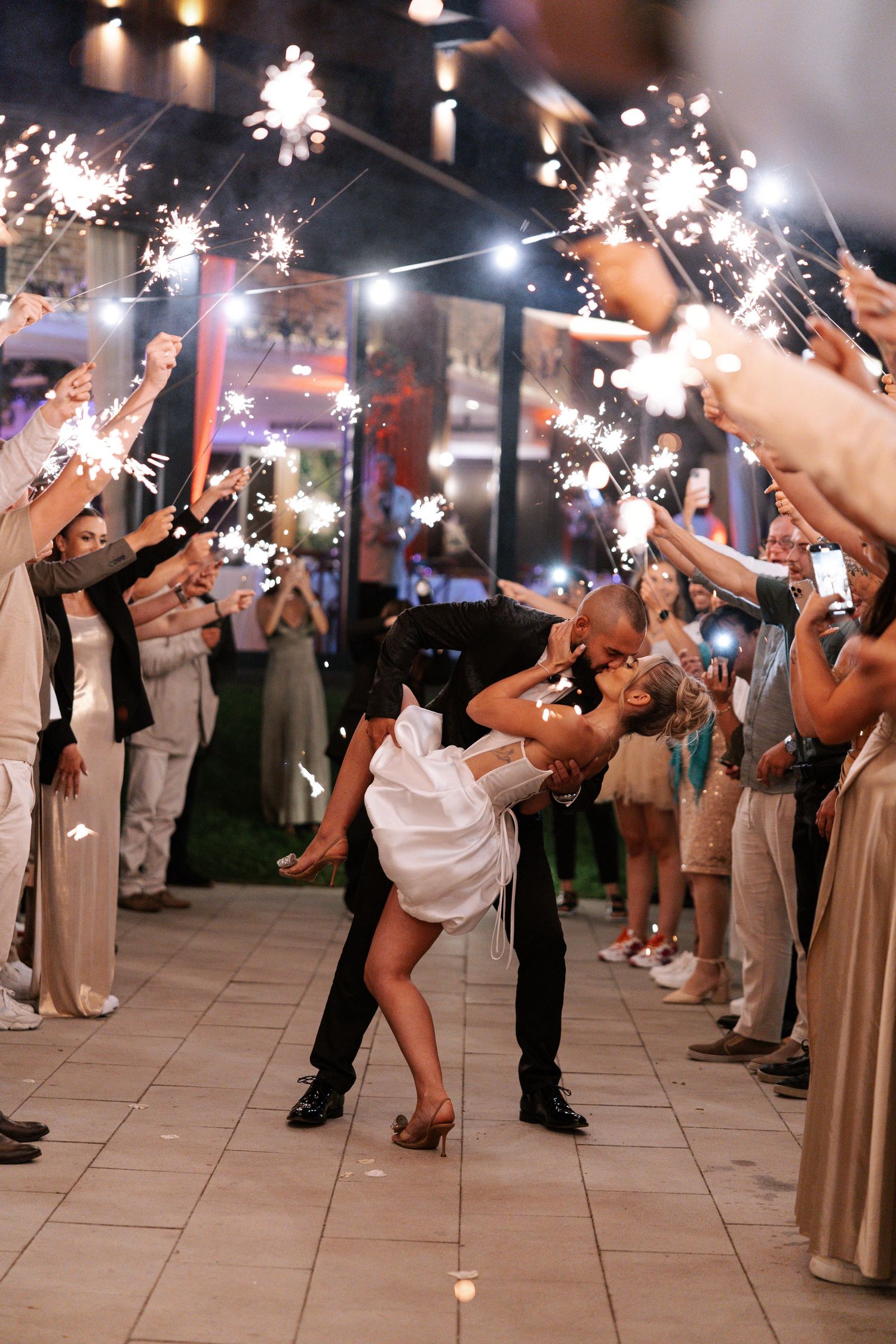 Romantic couple dips during sparkler exit at wedding reception while guests line walkway with sparklers.