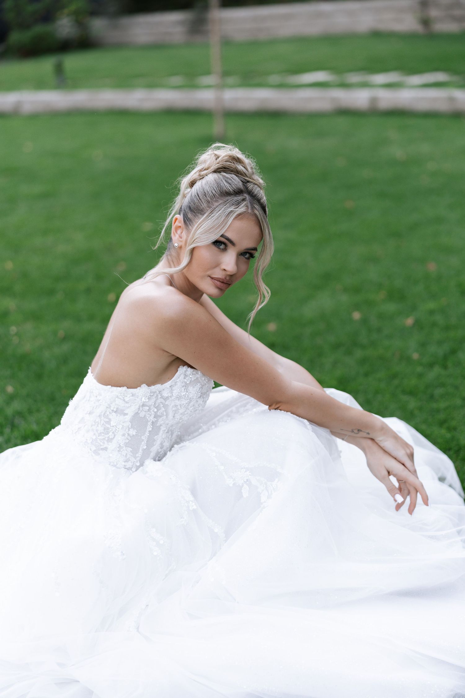 A bride in a white lace wedding gown poses gracefully on manicured grass for a romantic outdoor portrait session.