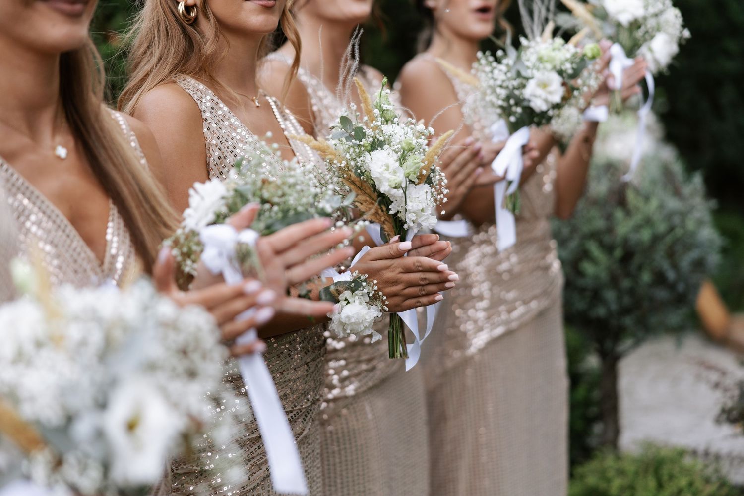 Wedding party members hold white and blush pink bouquets while wearing elegant sparkly dresses at an outdoor celebration.