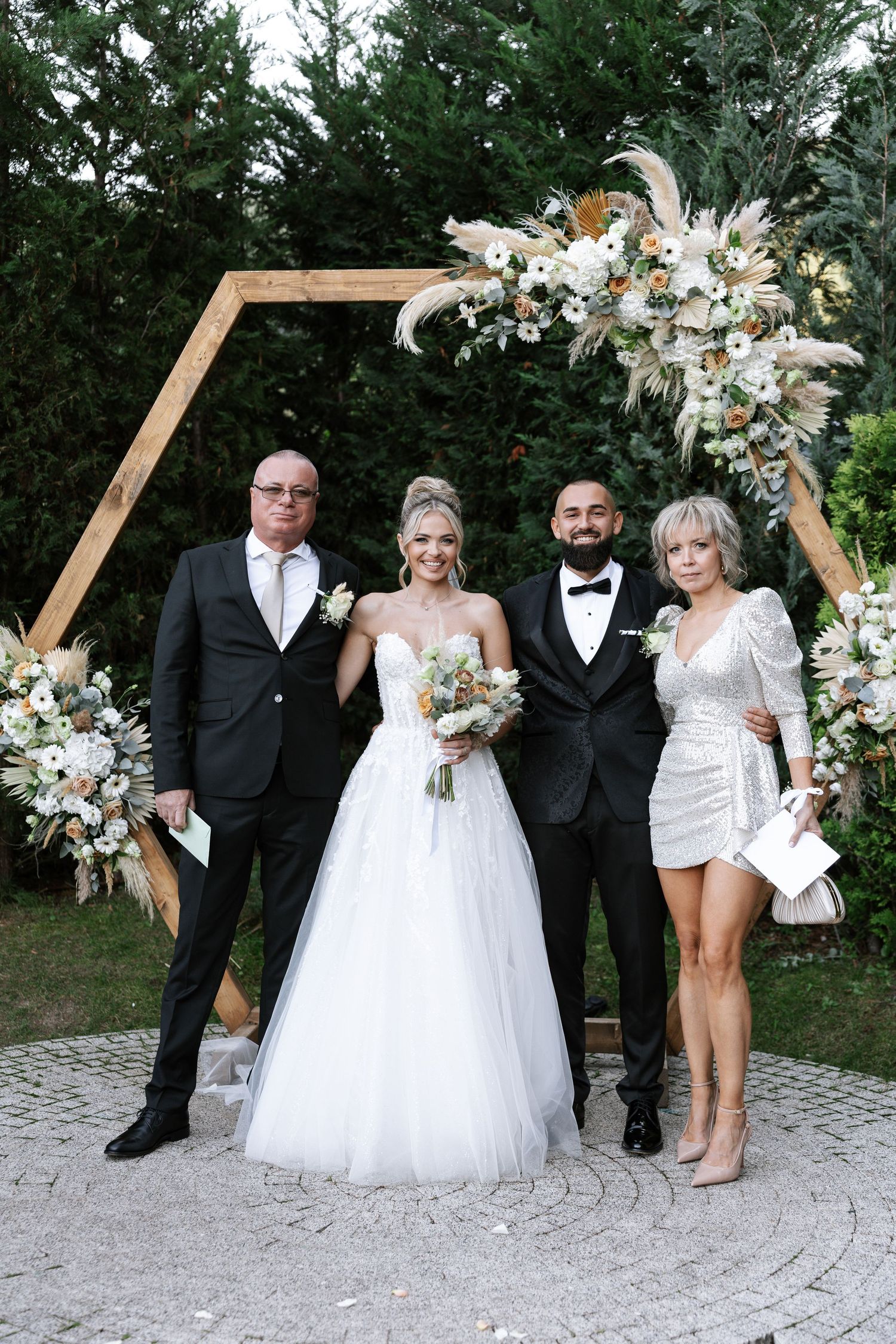 Wedding group poses under wooden hexagonal arch decorated with white and cream floral arrangements outdoors.