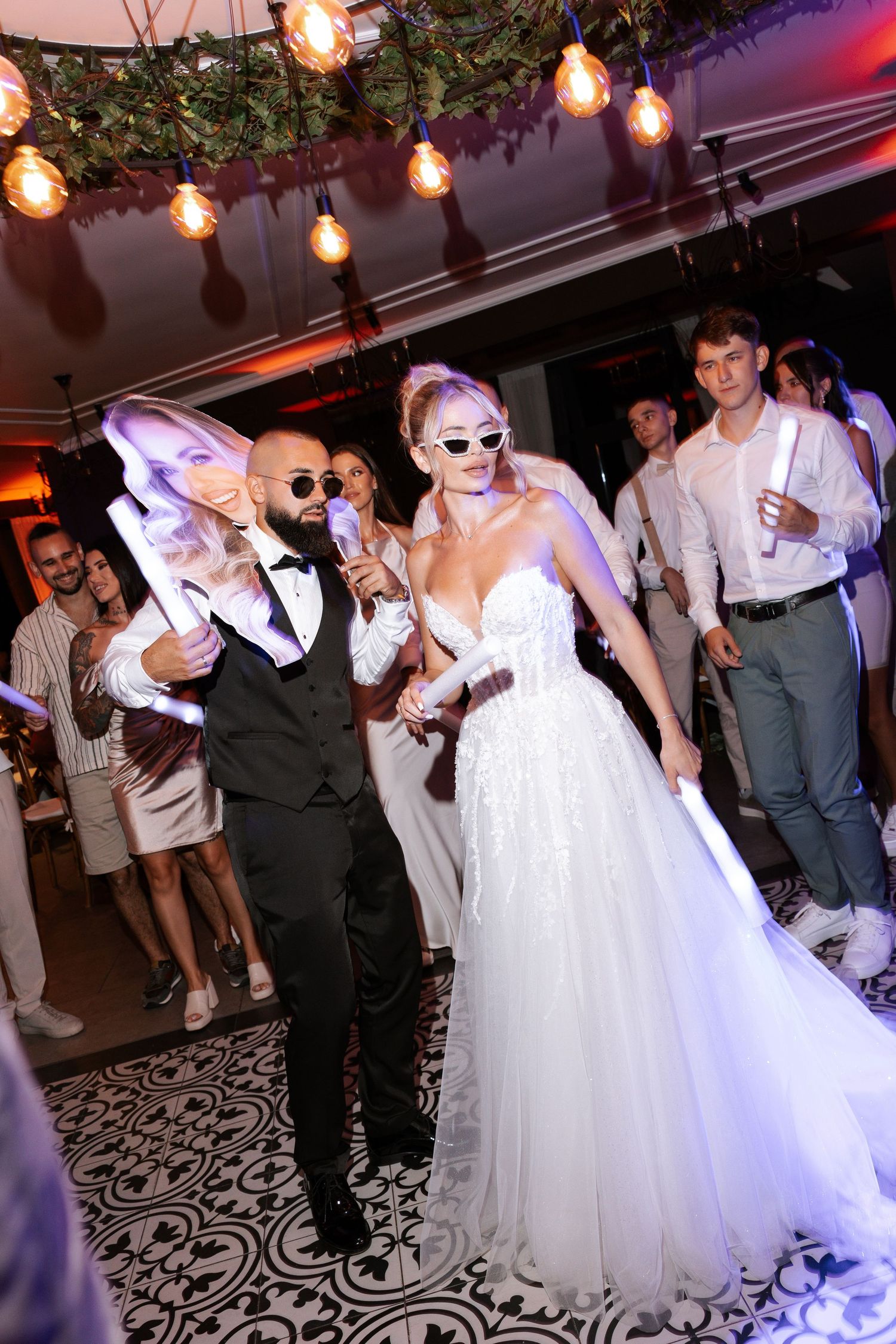 Wedding couple wearing sunglasses dance together under string lights on a patterned floor at their reception celebration.