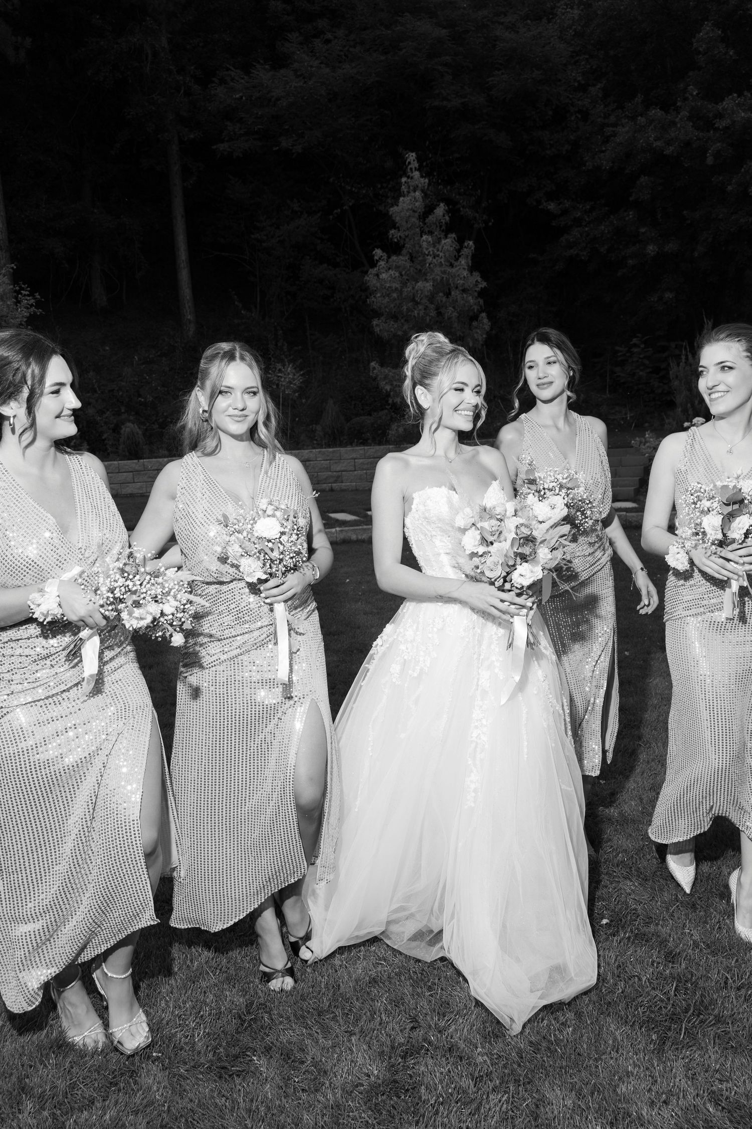 Black and white photo of a bridal party in sparkly dresses holding bouquets while standing on grass outdoors.