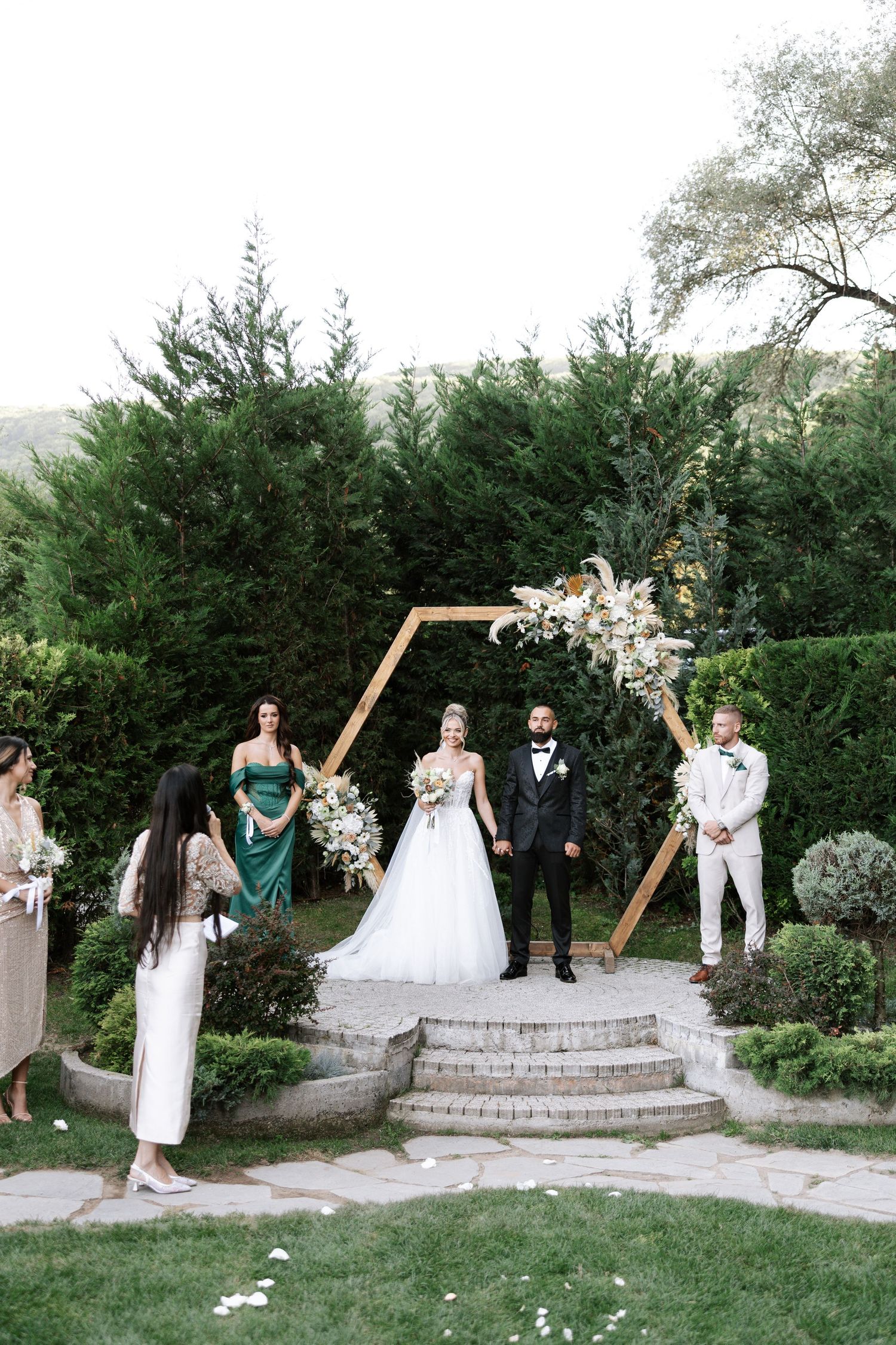 An outdoor wedding ceremony setup with a hexagonal wooden arch decorated with white flowers against a backdrop of evergreen trees.