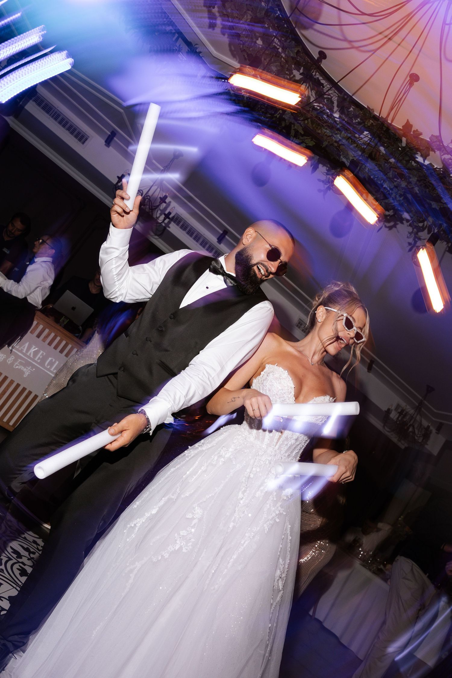 Wedding couple dancing with glowing LED foam sticks at their reception under colorful party lights.