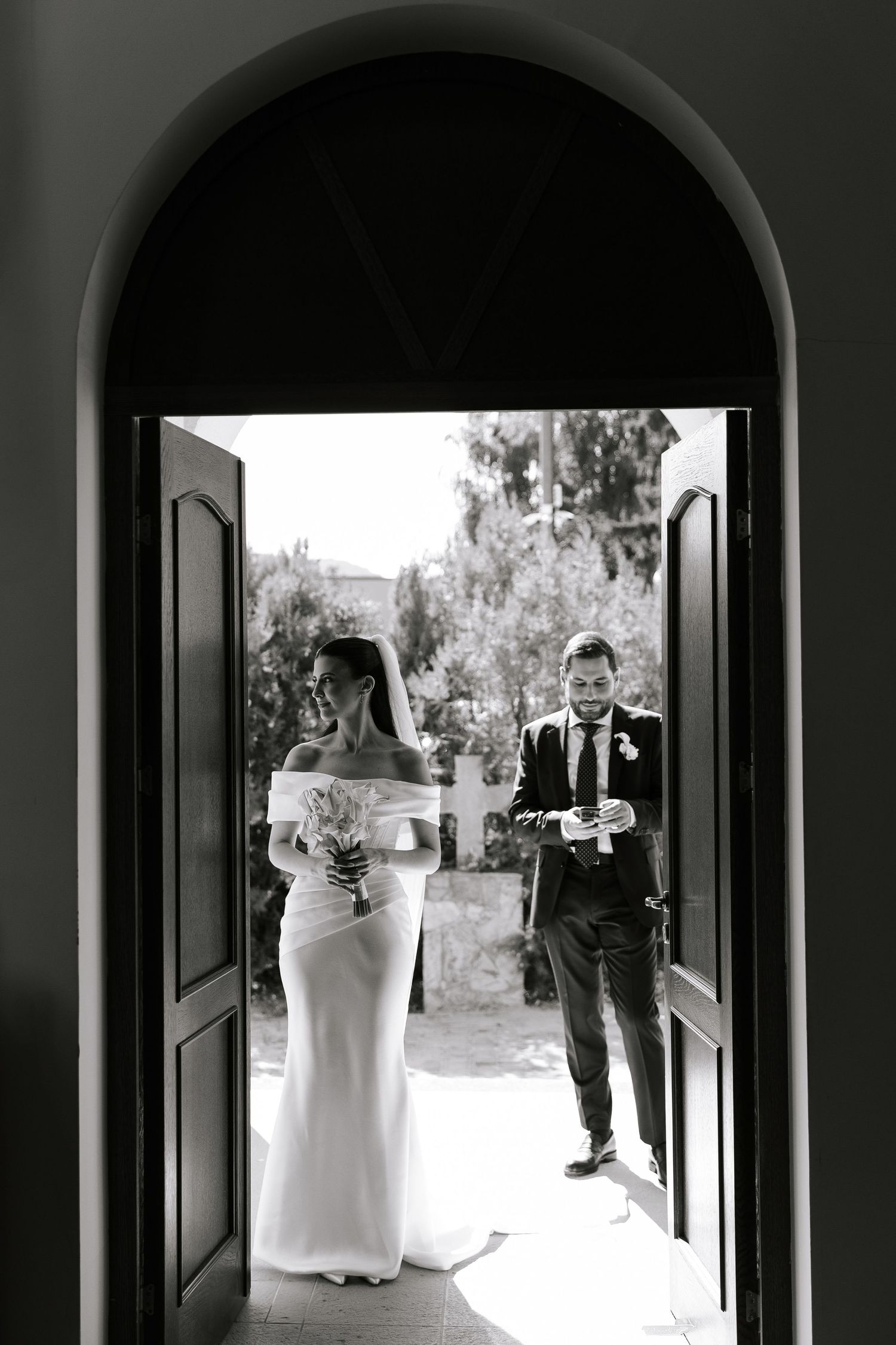 Black and white wedding photo showing a couple walking through an arched hallway framed by dramatic shadows.