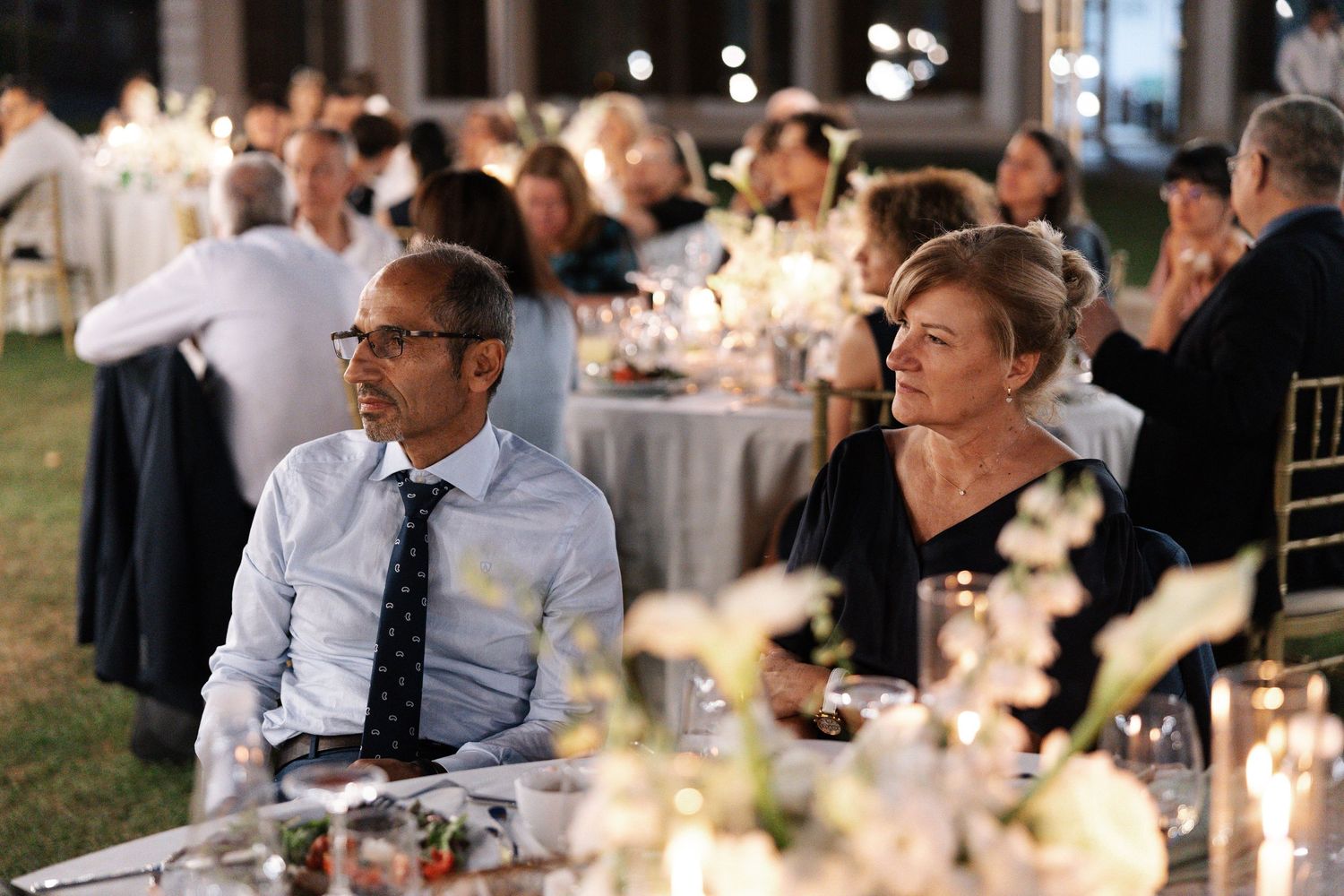 Wedding guests seated at elegant tables with glowing candles and floral centerpieces during an evening reception.