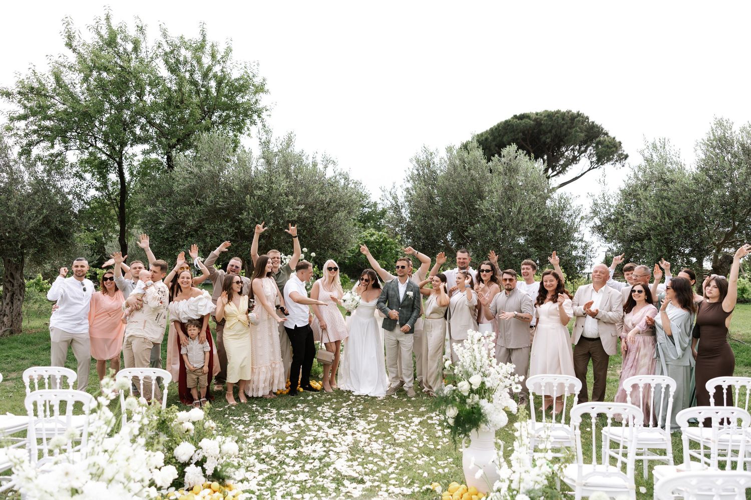 Large wedding party celebrates outdoors in an olive grove with white chairs and floral decorations.