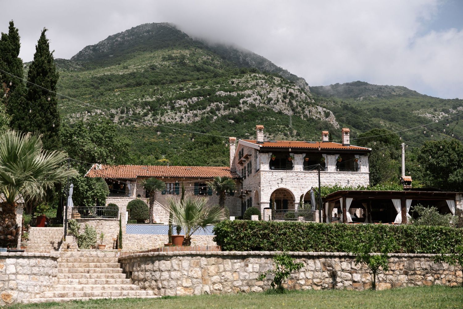 Mediterranean villa with white arches and red tile roof nestled against green mountainside in scenic landscape.