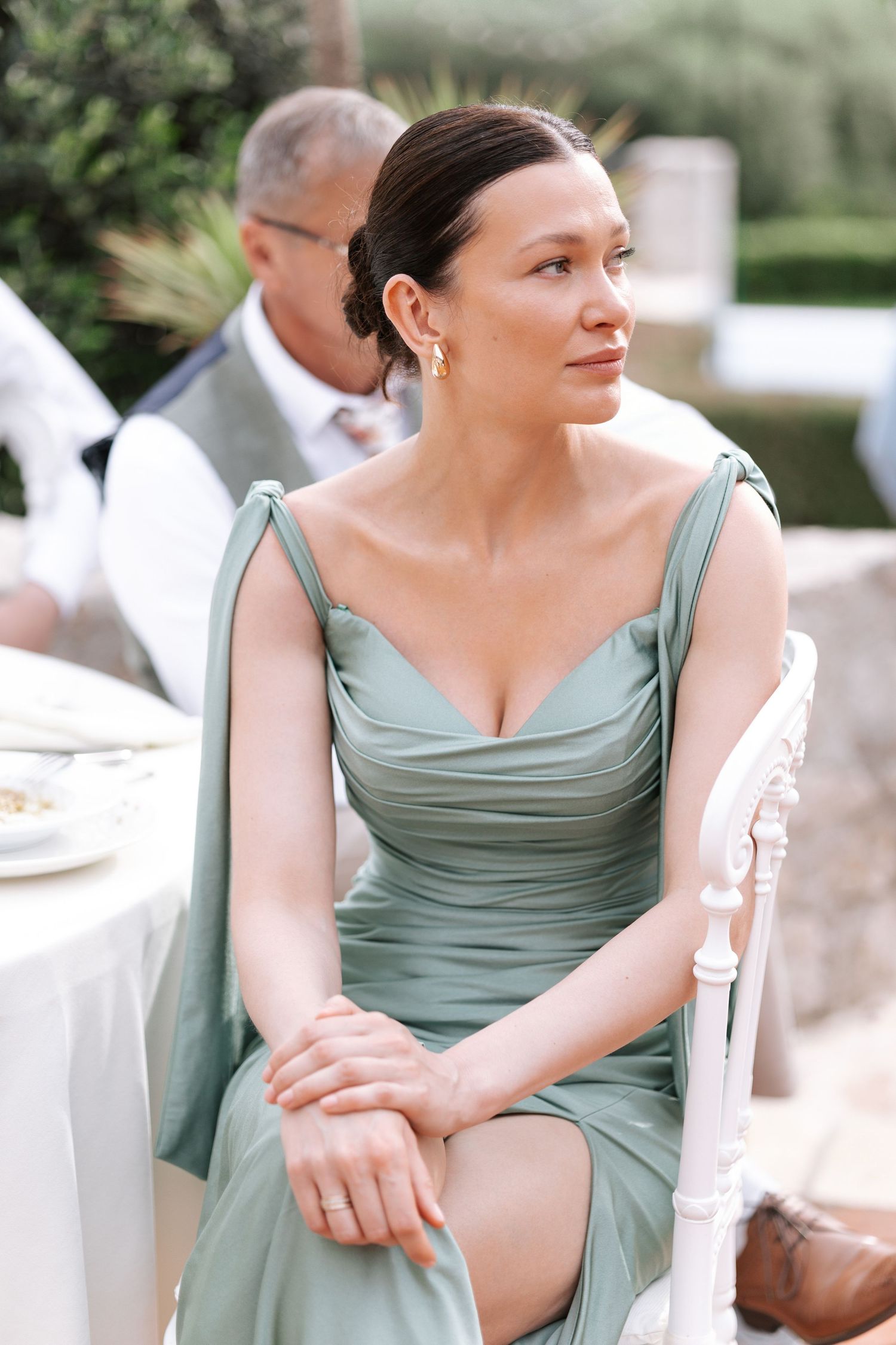 Woman in sage green bridesmaid dress sits elegantly at outdoor wedding reception.