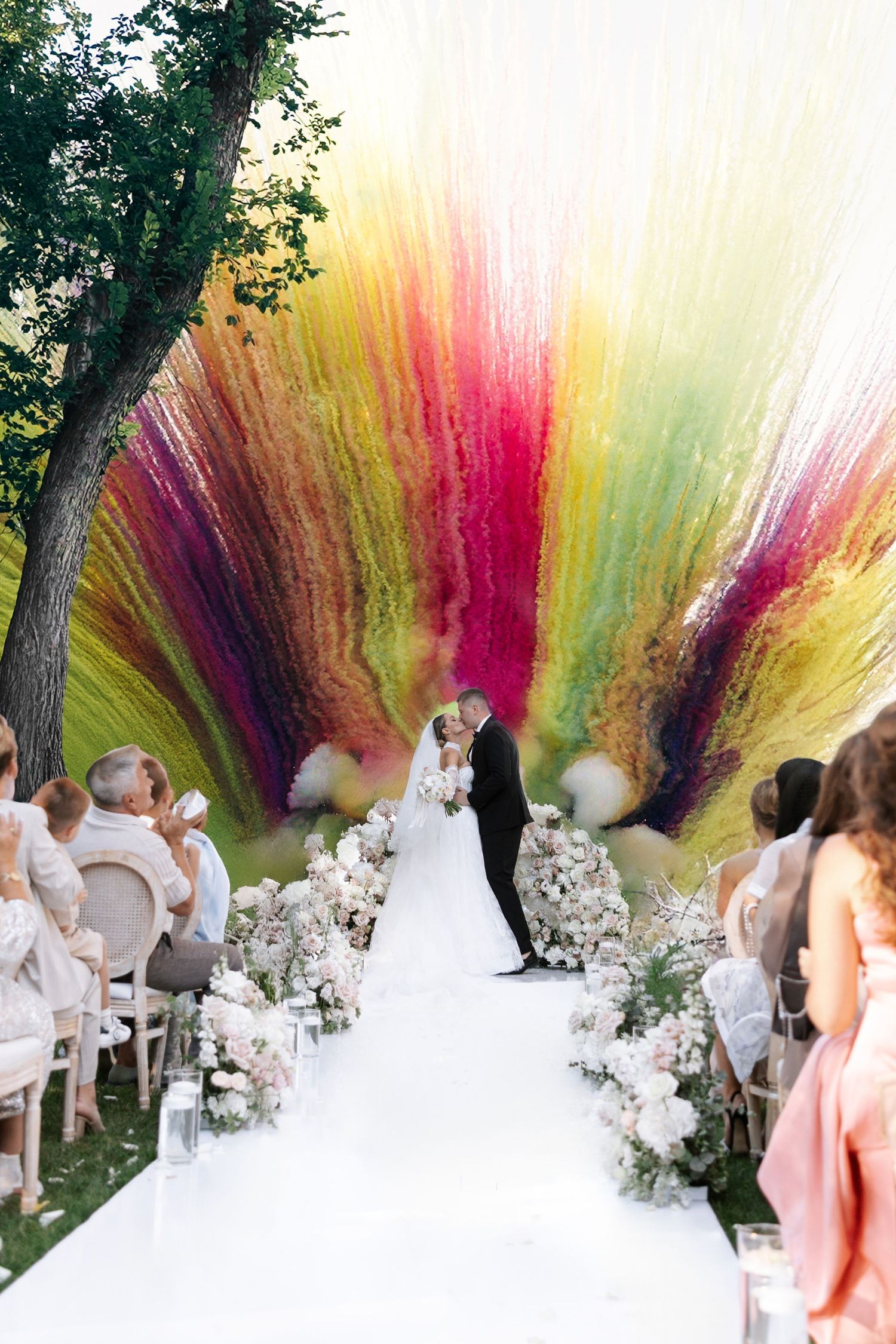 Vibrant rainbow smoke effect creates dramatic backdrop for outdoor wedding ceremony with white floral decorations.
