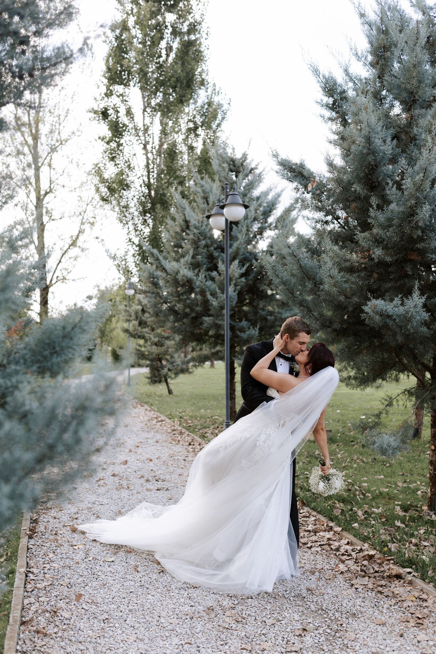 A romantic wedding portrait on a gravel path surrounded by evergreen trees.