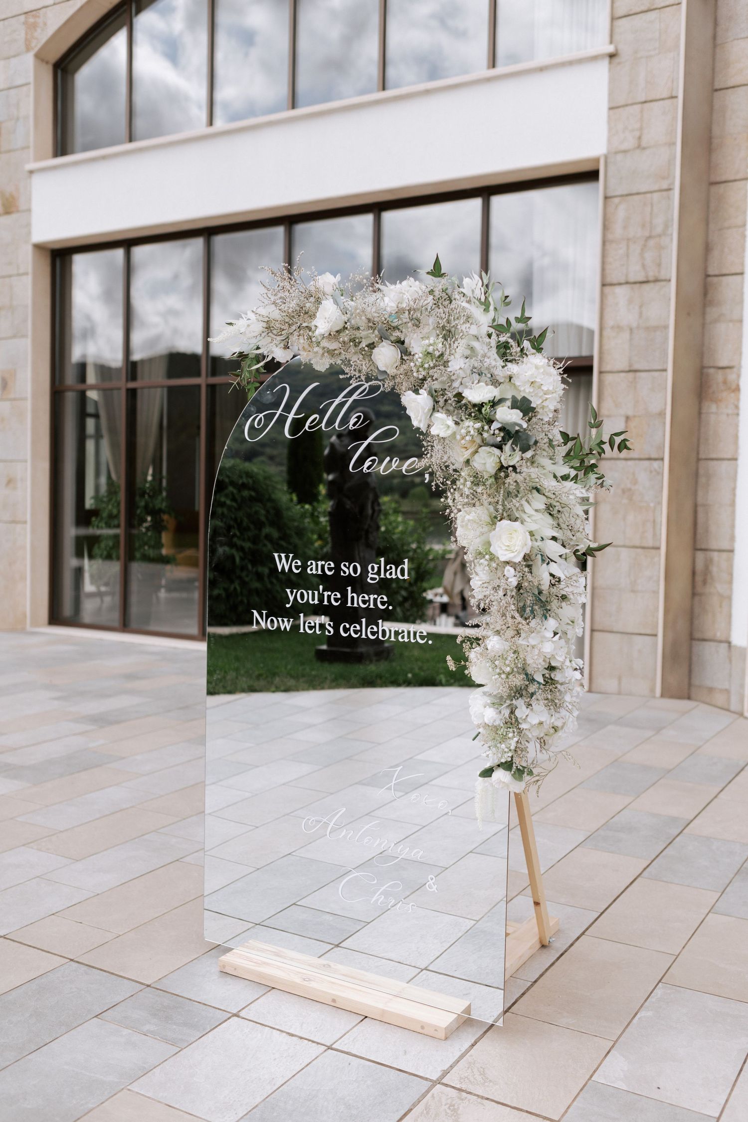 A mirror welcome sign decorated with white floral arrangements stands outside a modern building.