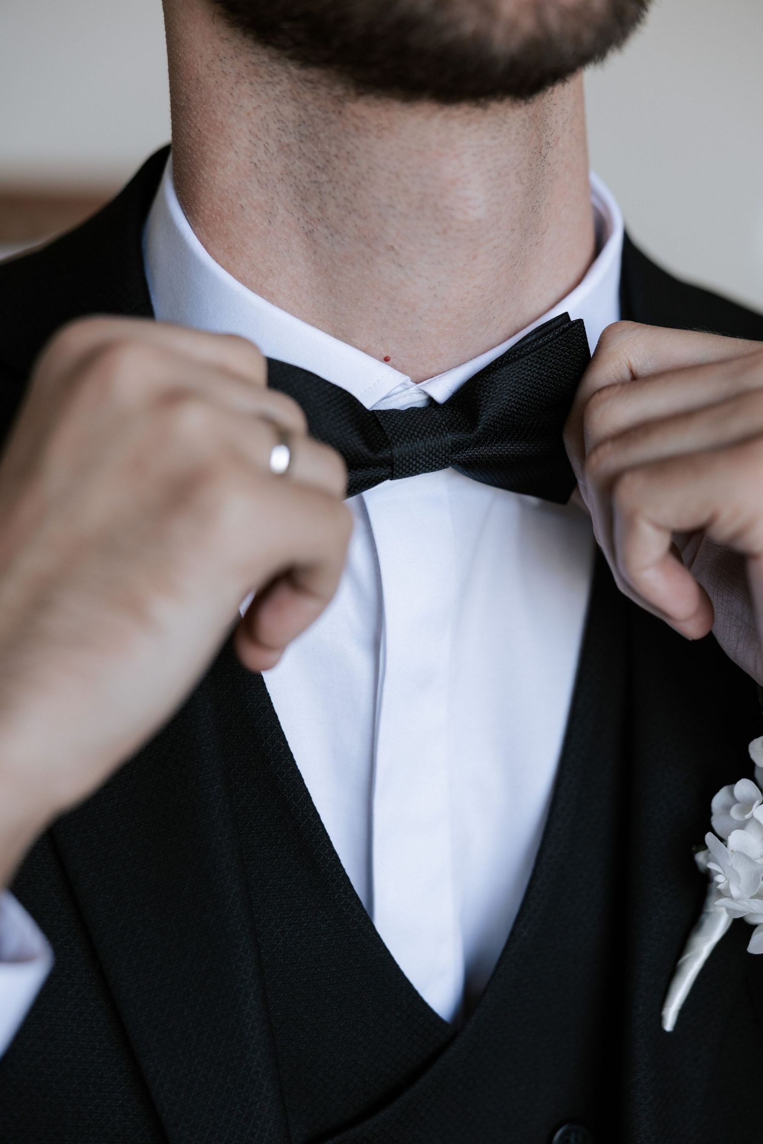 Close-up of hands adjusting black bowtie on formal tuxedo with white boutonniere.