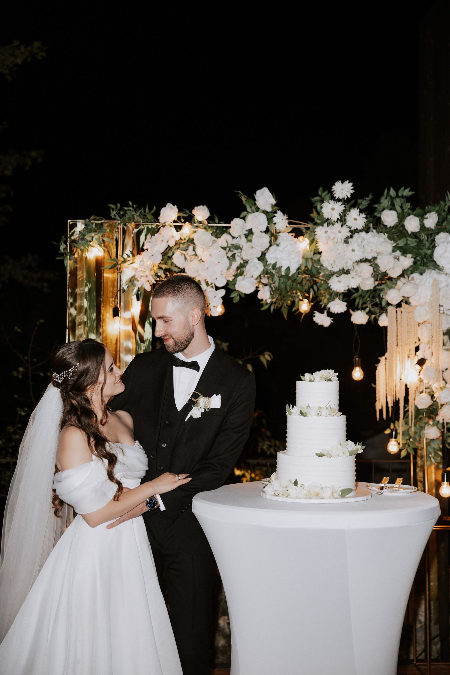 A tender moment between newlyweds near their wedding cake decorated with white flowers and twinkling lights.