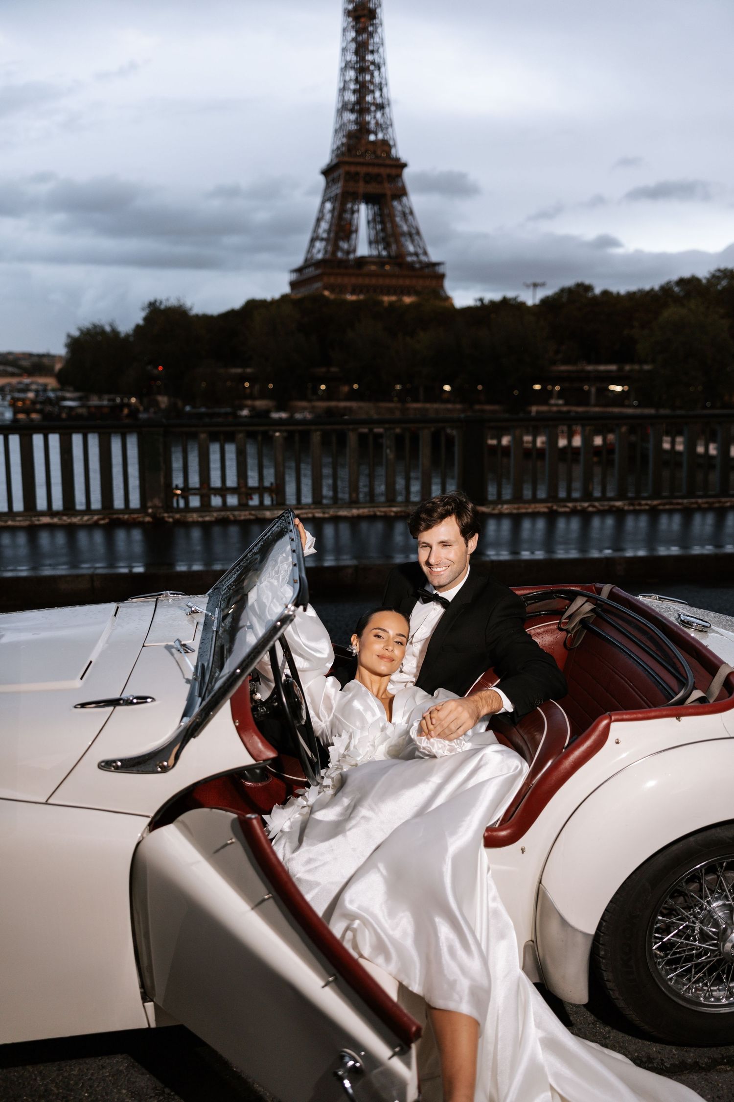 A couple poses in a vintage convertible car with the iconic Eiffel Tower in the background at dusk.