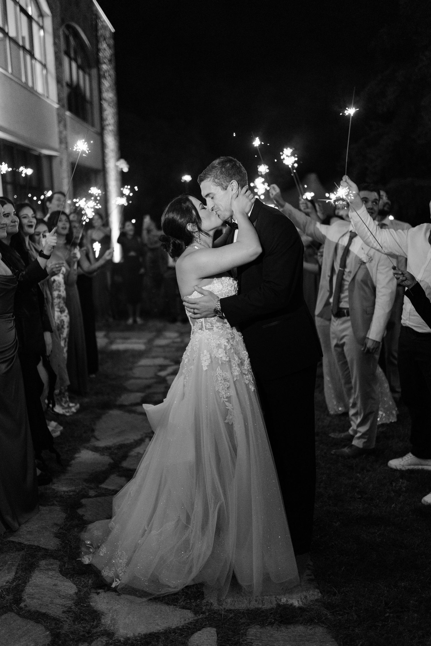 Romantic sparkler exit with couple embracing while guests hold sparklers in black and white.Wedding photographer Bulgaria