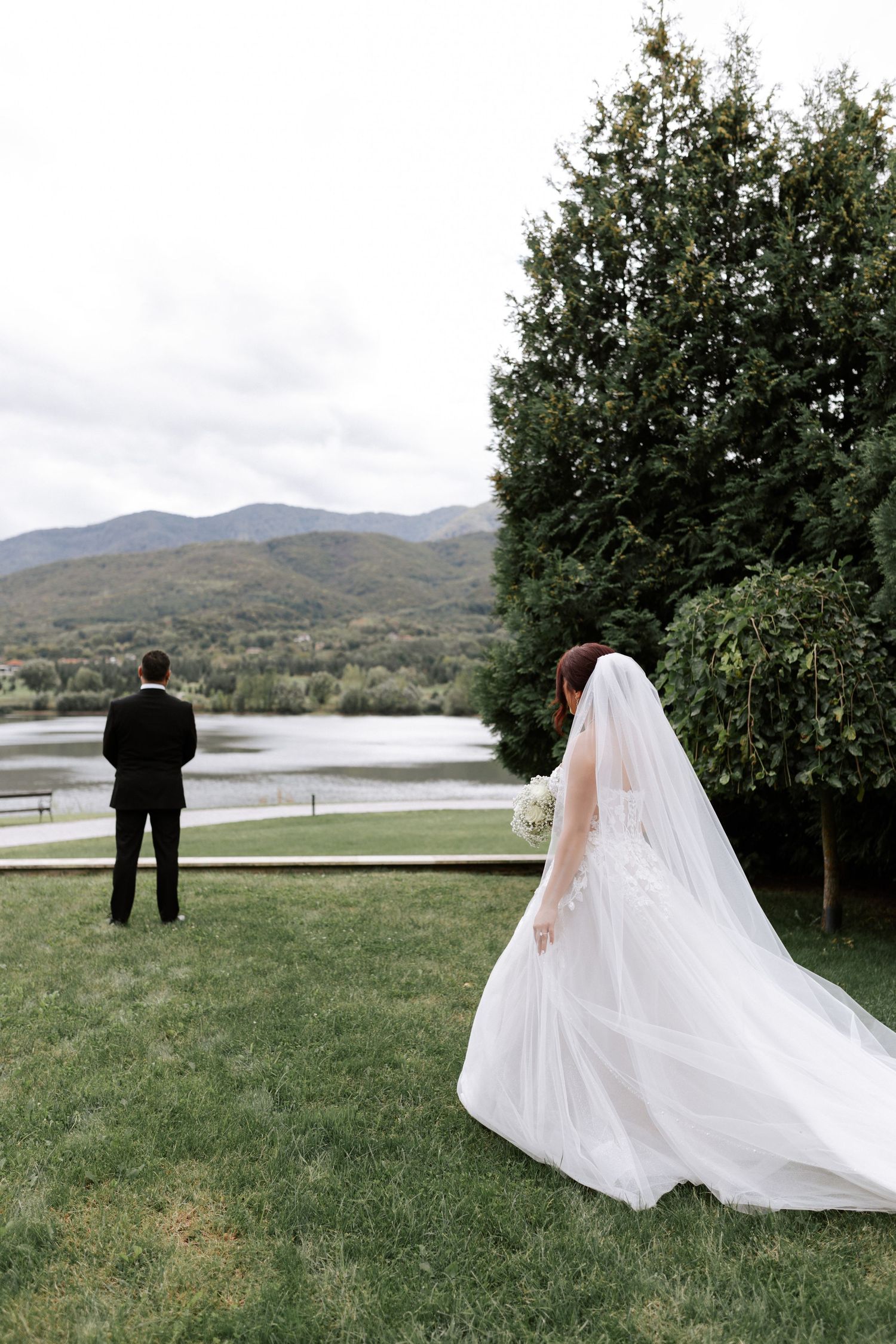Bride and groom share first look moment on a lawn with mountain views and lake backdrop.