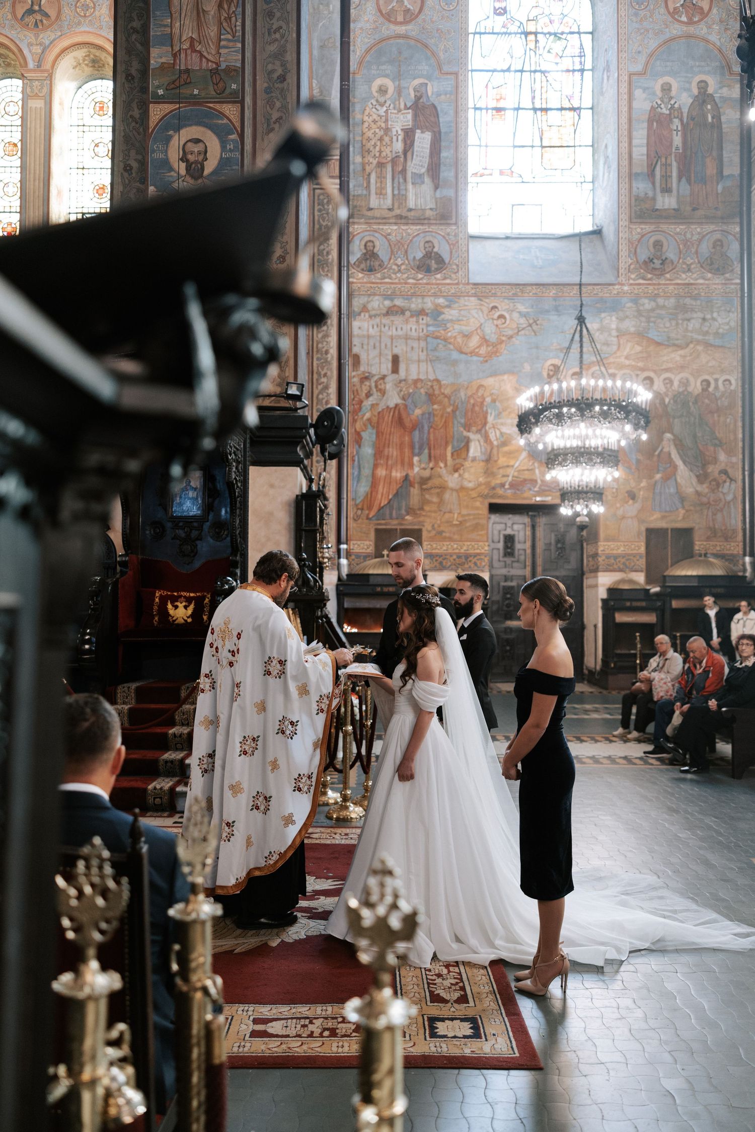 Orthodox wedding ceremony in progress inside historic church with painted walls and chandeliers.