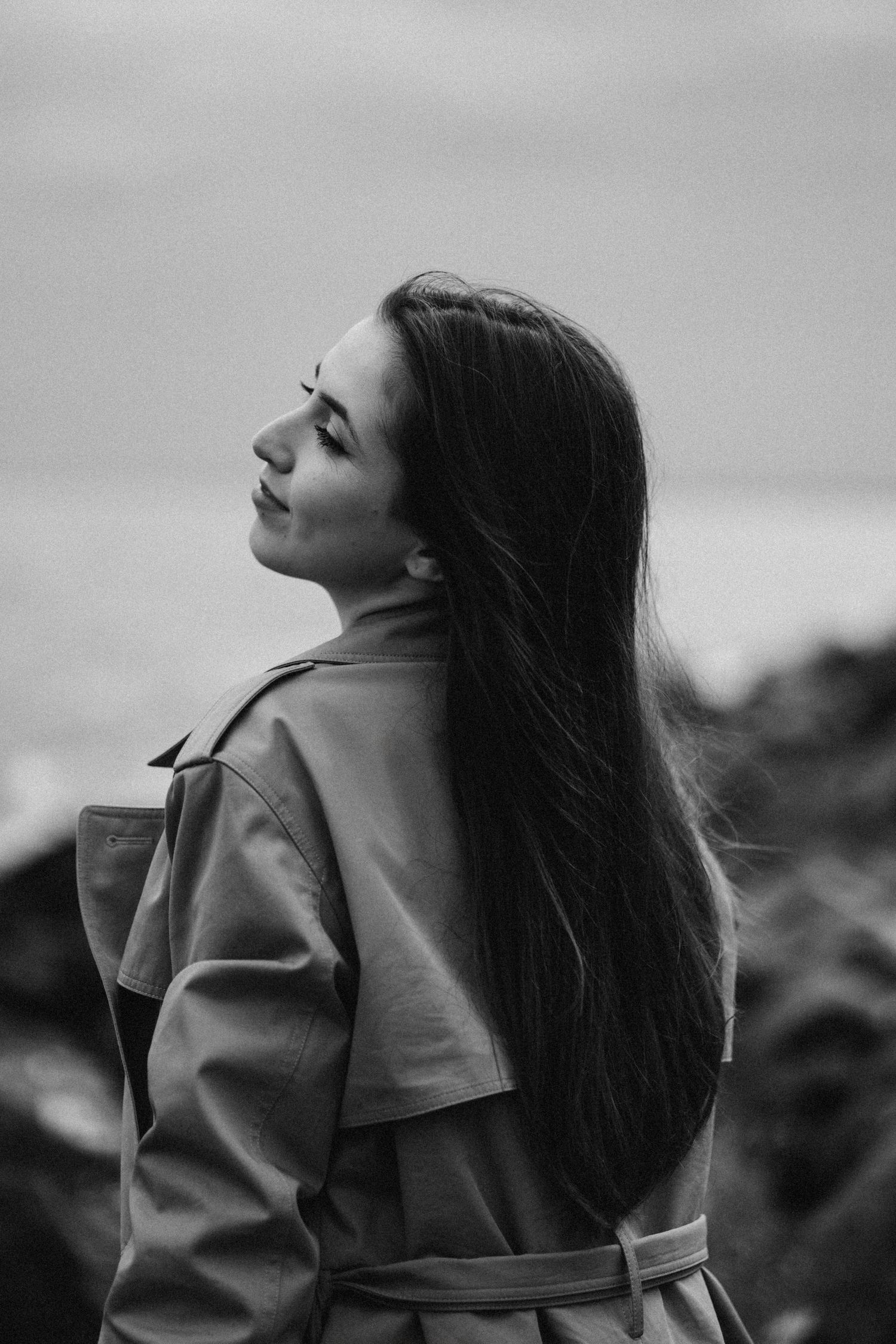 Woman in trench coat poses for an artistic black and white portrait with long flowing hair.