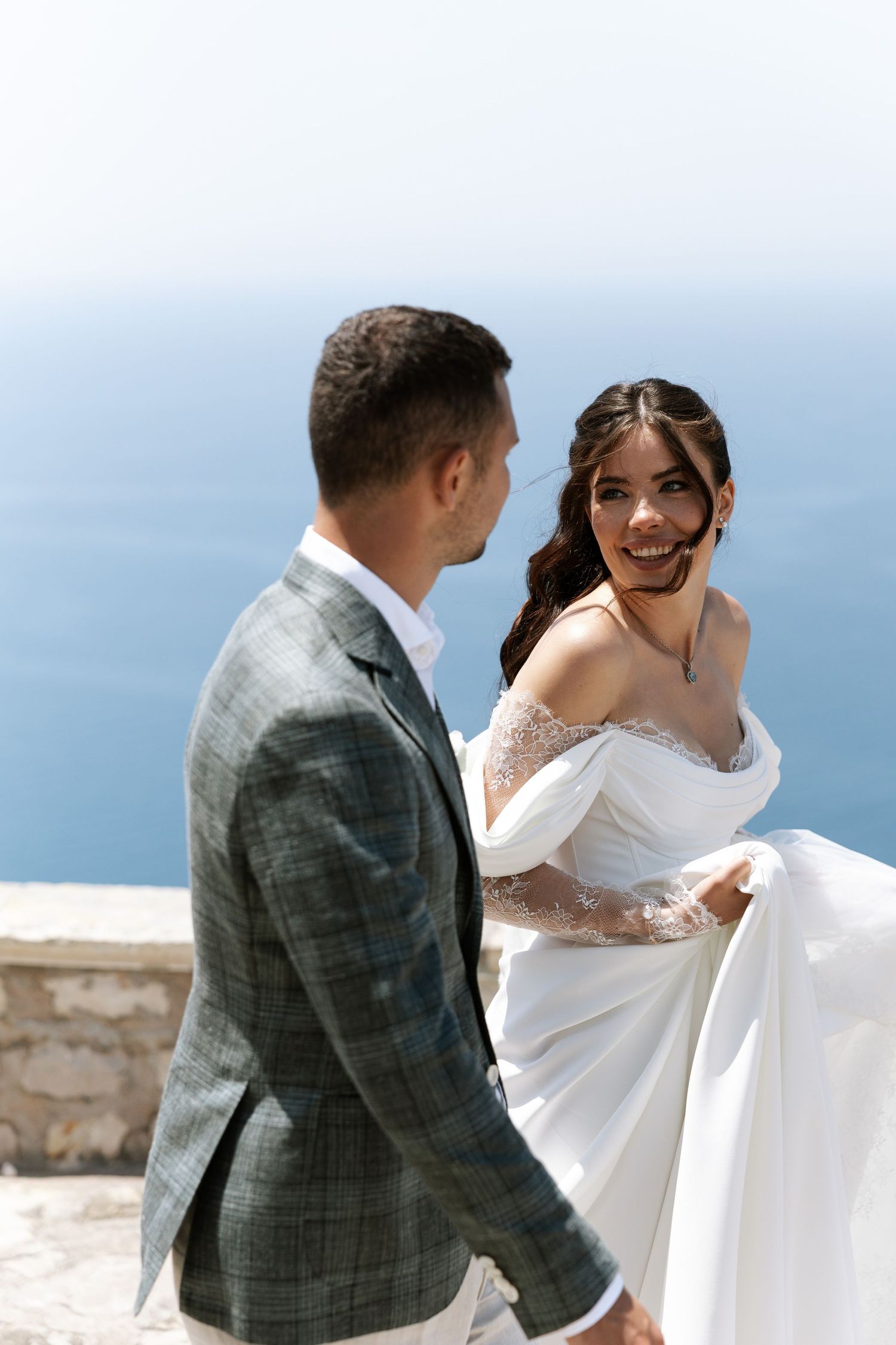A couple in wedding attire shares a joyful dance moment on a scenic overlook by the ocean.