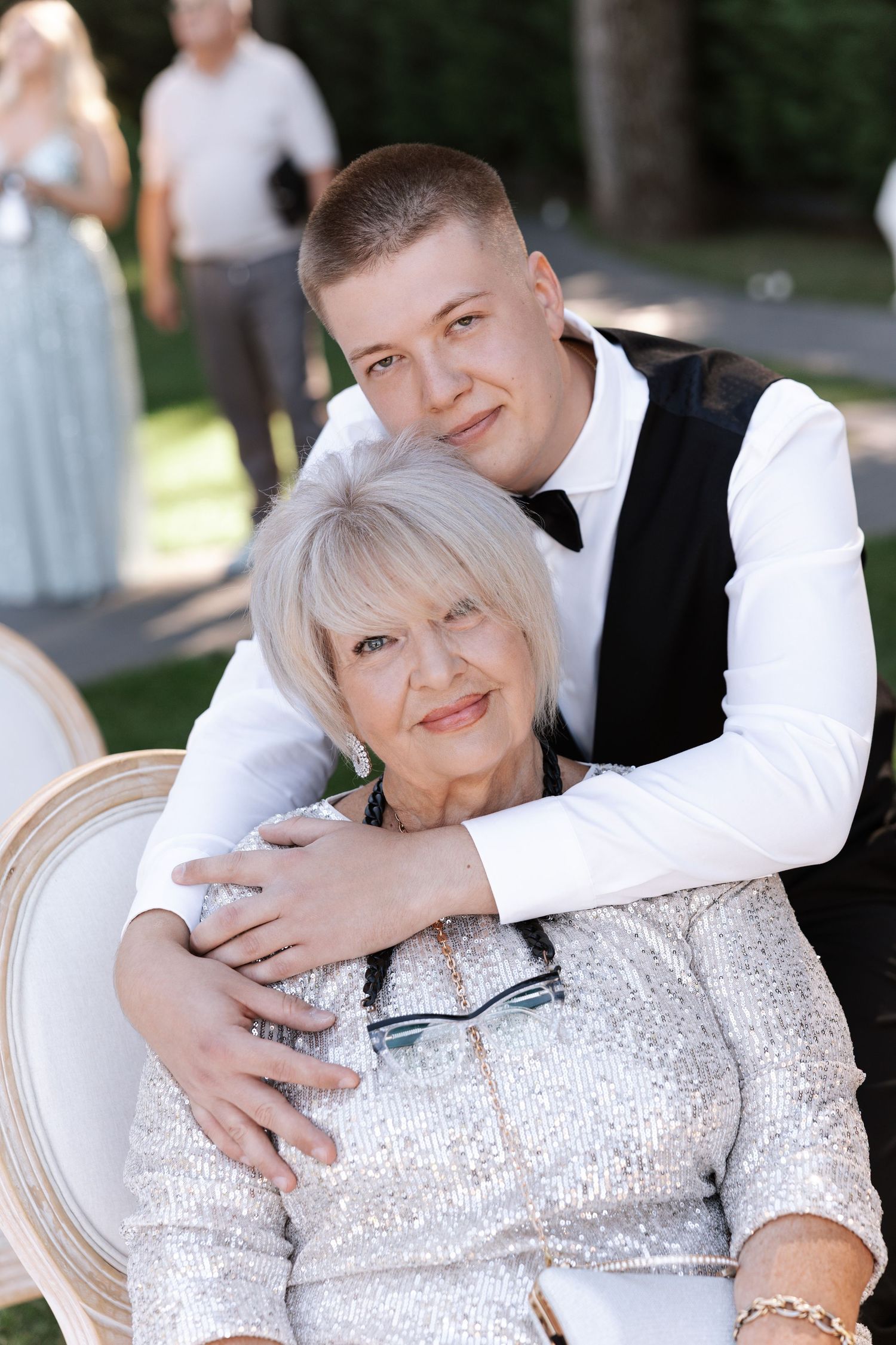Two wedding guests share a warm embrace while seated in an elegant chair outdoors.