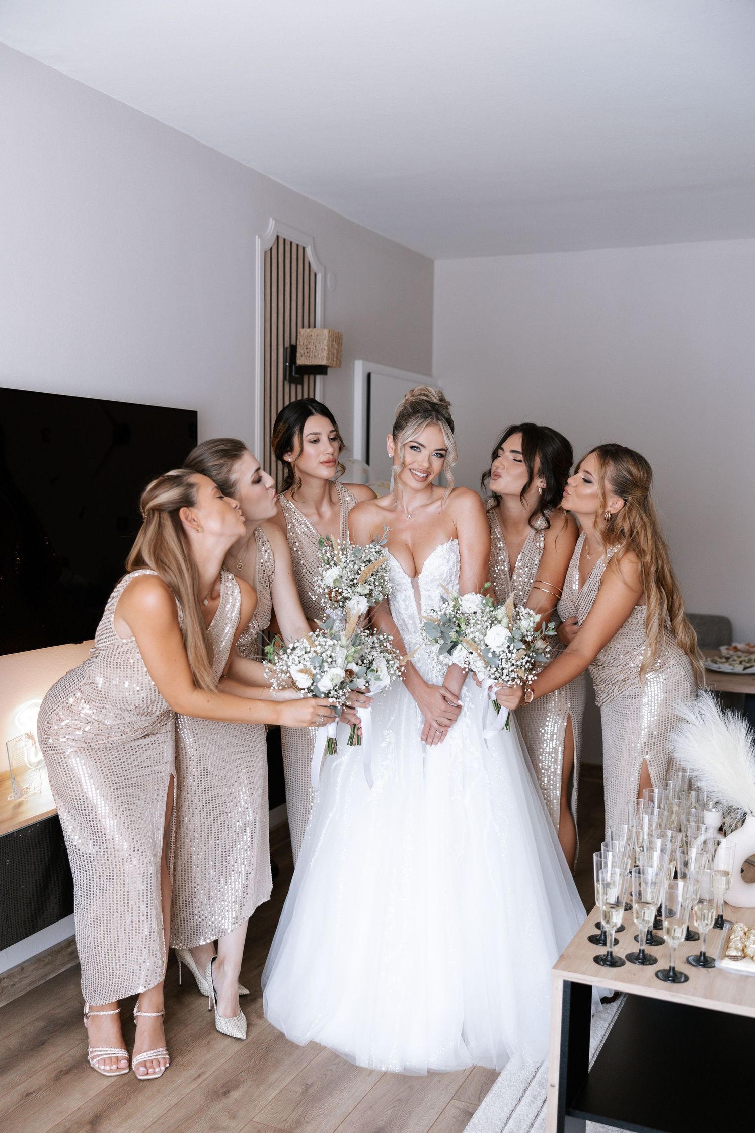 Bridesmaids in sparkly pink dresses gather around bride in white gown for candid wedding party photos.