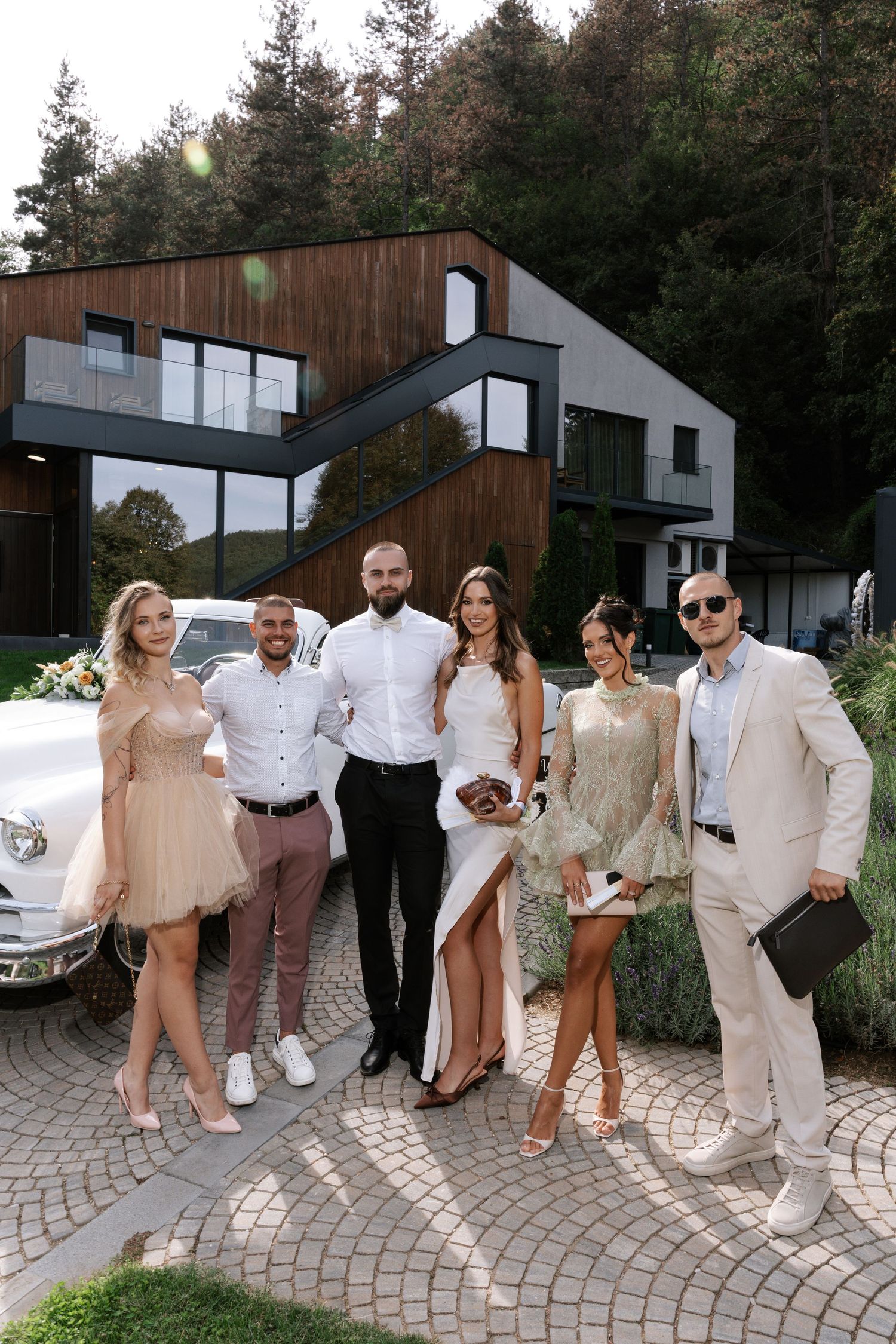 A group of well-dressed guests pose together on a stone paved driveway in front of a modern luxury house with wood accents.